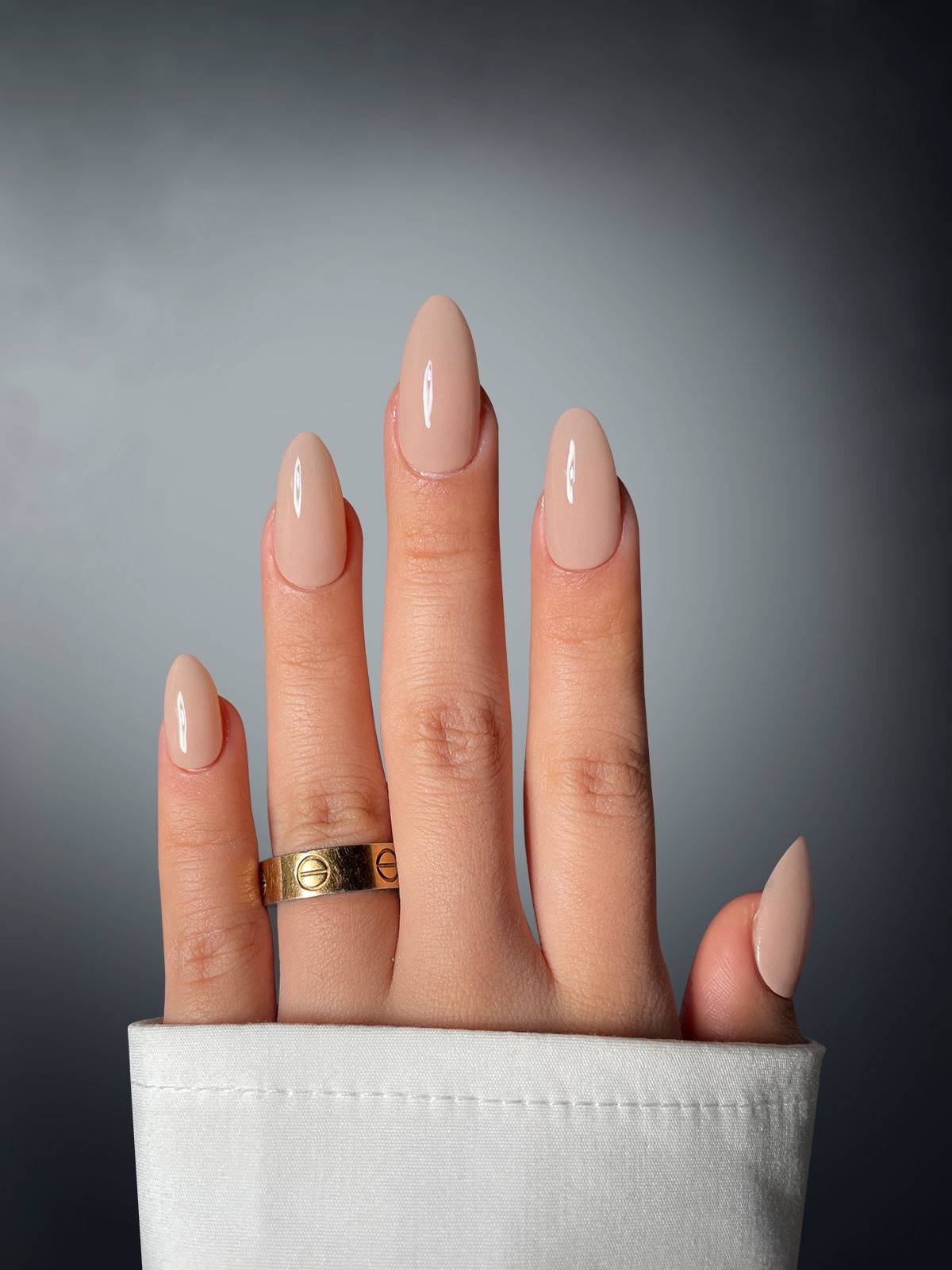 A hand with neatly manicured, glossy nude nails features Sassy Saints' Cappuccino Chic Dipping Powder. Against a gray backdrop, the person wears a gold ring on their middle finger and sports a white sleeve.