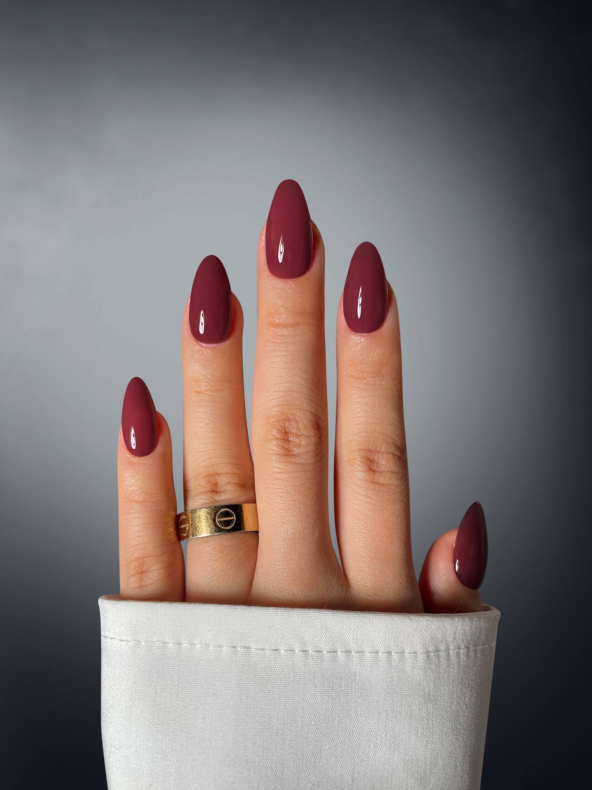 A hand with long nails polished in Sassy Saints’ Merlot Moxie Dipping Powder and a gold ring rests on a gray background, partially covered by a white sleeve.