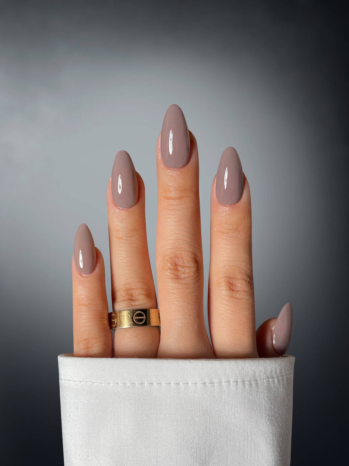 A hand with four glossy taupe nails, featuring Sassy Saints - Brings The Salon Home Beguiling Bombshell Dipping Powder, extends from a white cuff. A gold ring adorns the middle finger against a dark-to-light gray gradient background.