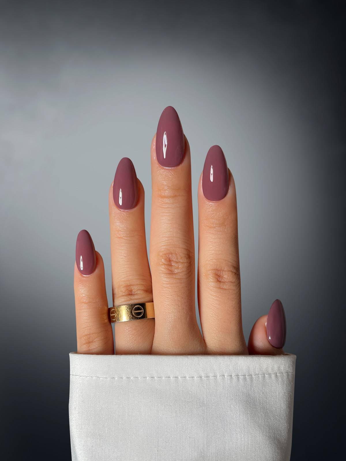 A hand displays Sassy Saints’ Graceful Goddess Dipping Powder in glossy mauve on four fingers, with a gold ring on the middle finger. The hand emerges from a white sleeve against a gradient gray background.