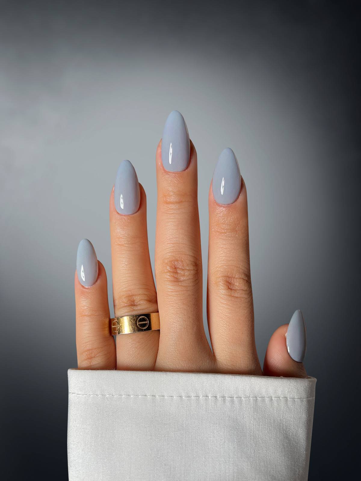 A hand with almond-shaped nails in Fog Fume Dipping Powder by Sassy Saints is shown against a gradient grey background, wearing a gold ring and a white long-sleeve shirt.