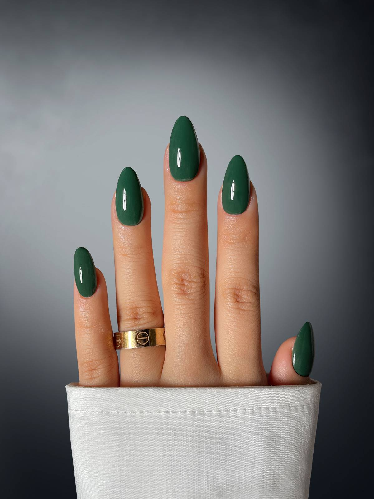 A hand with green oval-shaped nails in Viridian Vogue Dipping Powder by Sassy Saints – Brings The Salon Home – wears a gold ring and emerges from a white sleeve, with fingers dramatically bent backward against a gradient gray background.