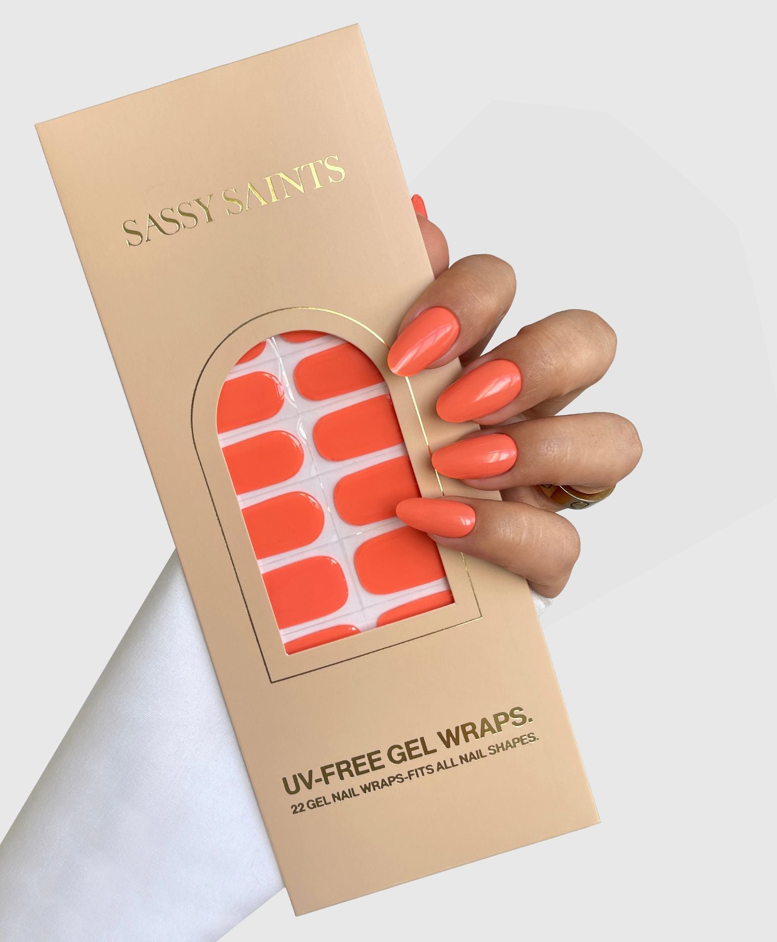 A hand with long, glossy, orange nails holds a Sassy Saints - Brings The Salon Home Papaya Glow Gel Wrap package in a matching bright orange shade against a neutral background.