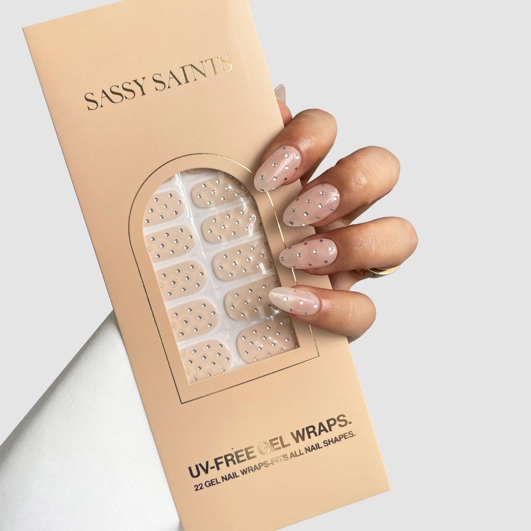 A manicured hand holds a Sassy Saints – Brings The Salon Home Platinum Spotlight Gel Wrap box, with nude-colored gel wraps featuring small silver dots visible through a window on the packaging.