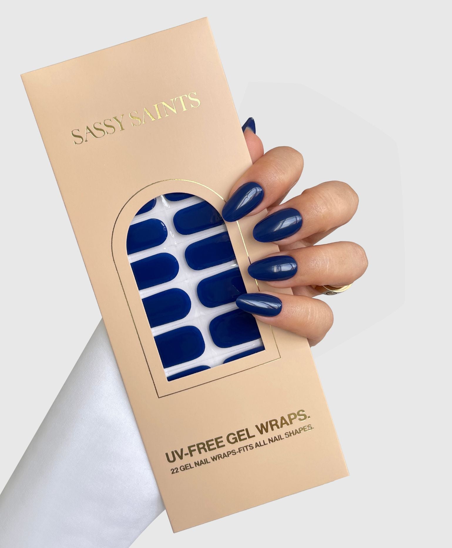 A hand with long, glossy Blue Eclipse Gel Wrap nails holds a beige Sassy Saints - Brings The Salon Home box showing matching blue gel nail wraps inside, against a light gray background.