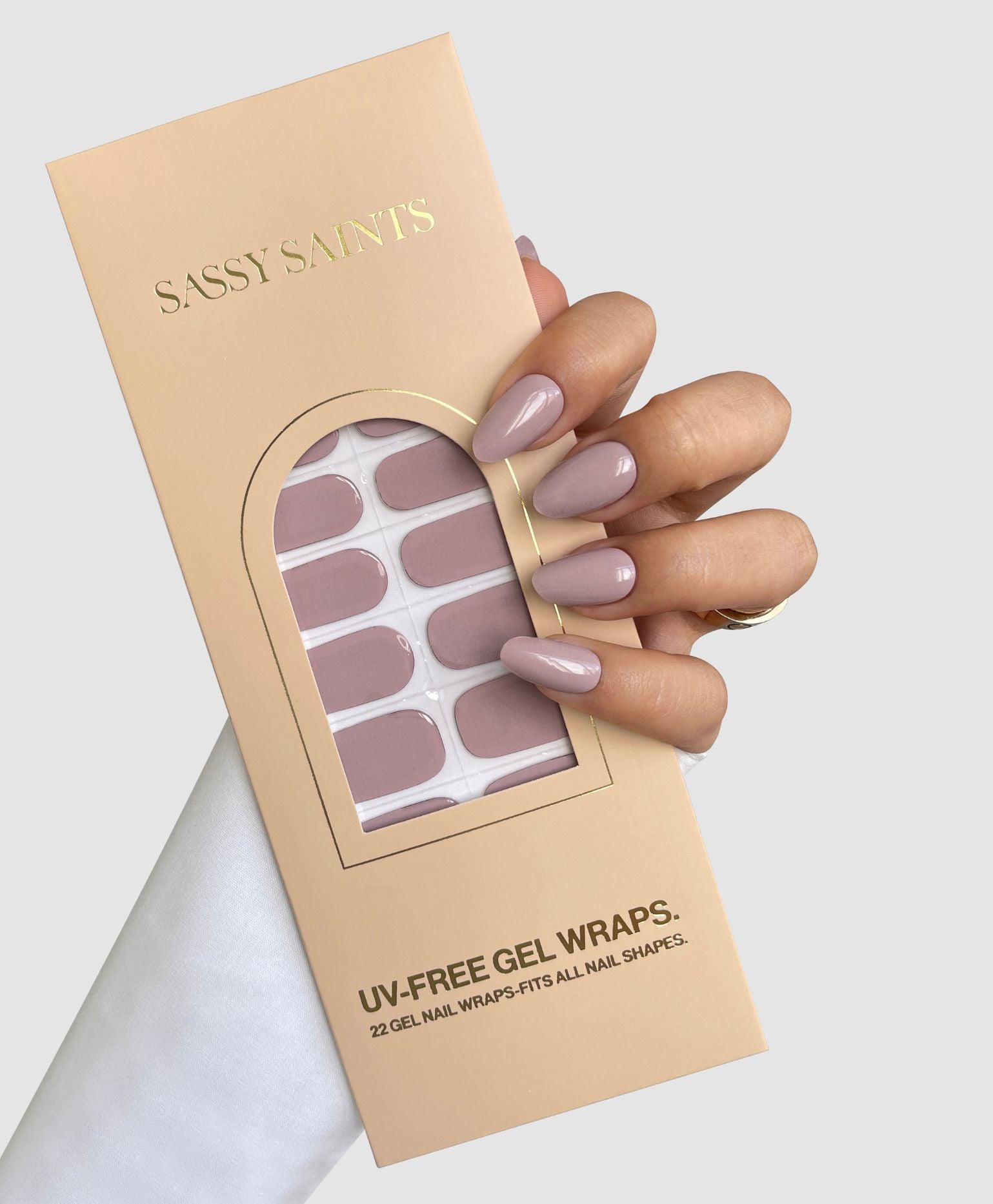 A hand with long, glossy nude-pink nails holds a box of Sassy Saints - Brings The Salon Home Neutral Flow Gel Wraps in a matching shade. The beige box features gold lettering and displays 22 UV-free gel wraps inside.