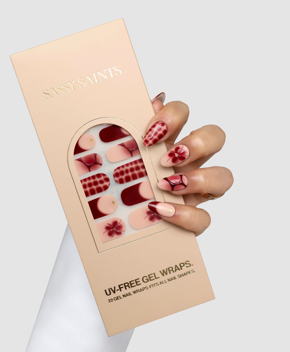 A hand with decorated nails holds a Sassy Saints - Brings The Salon Home Burgundy Bloom (3D) Gel Wrap box. The gel nail wraps feature matching red, pink, and floral designs. The background is plain light grey.