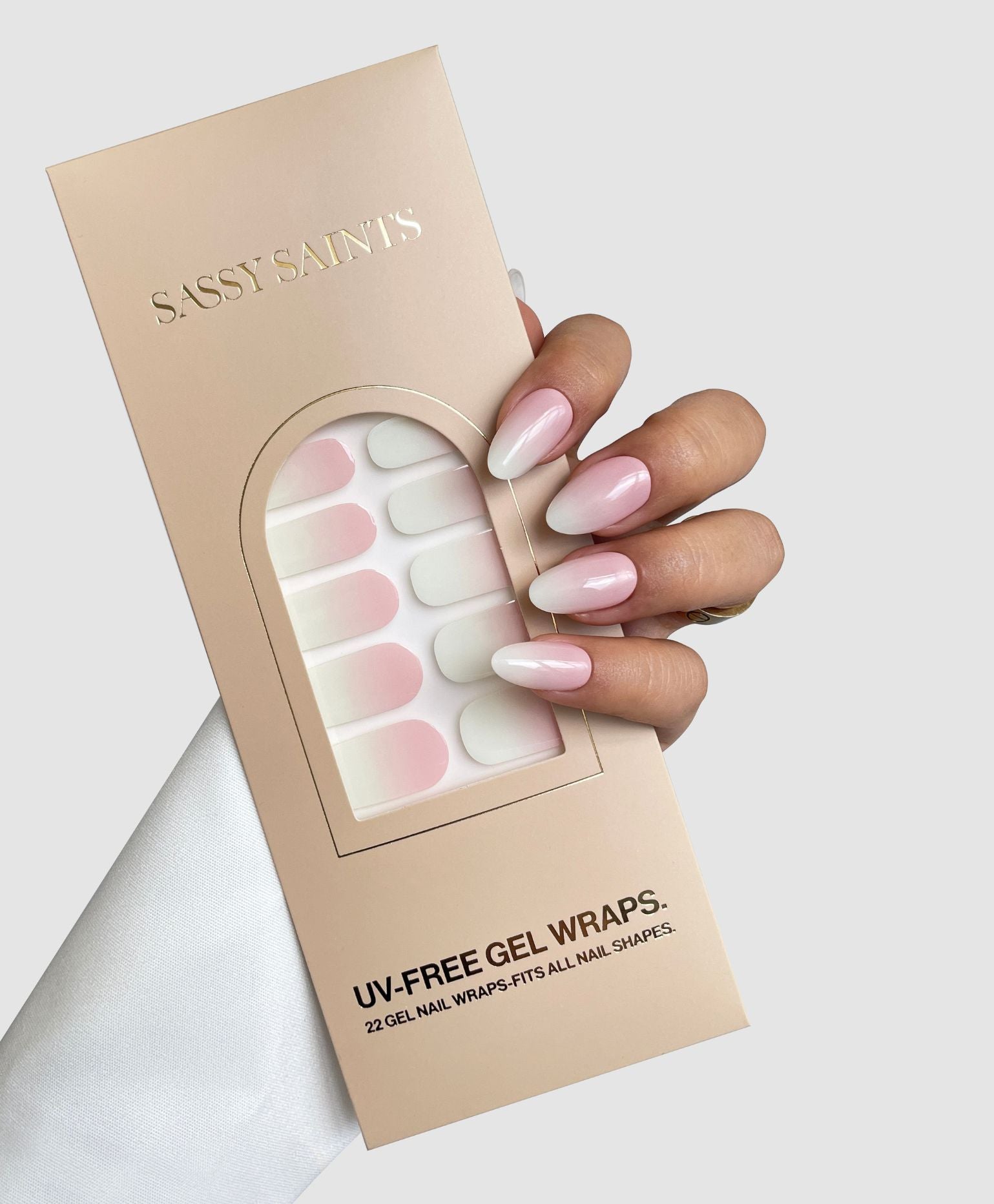 A hand with pink ombré gel nails holds a box of Sassy Saints - Brings The Salon Home Classic Ombre Gel Wraps in matching pink and white shades, set against a light gray background.