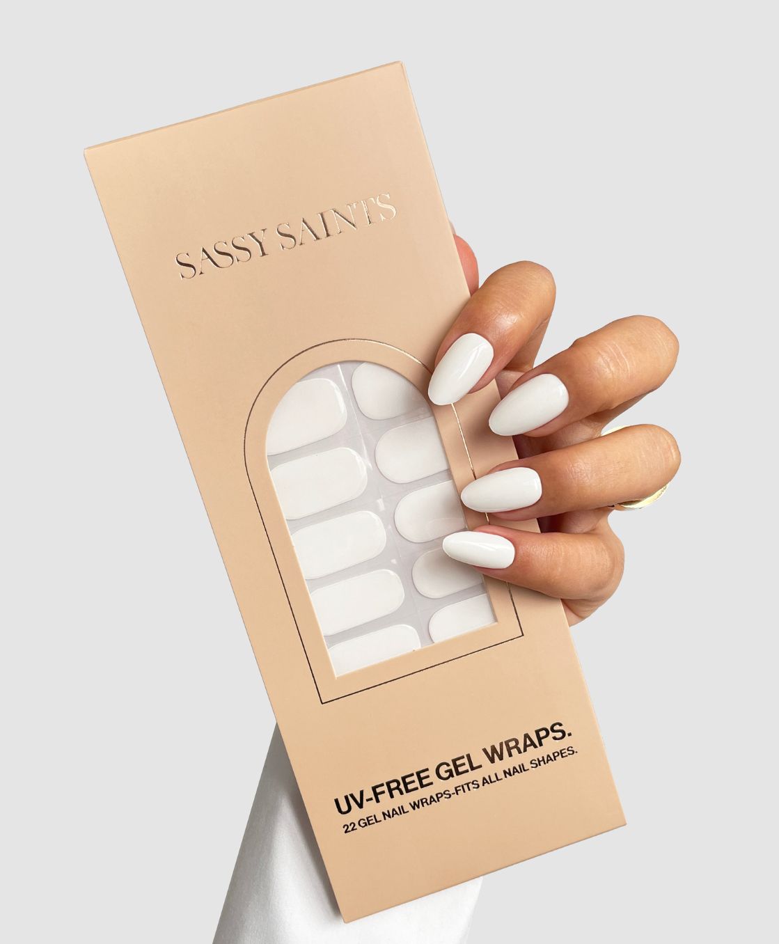 A hand with long, white, glossy nails holds a Sassy Saints - Brings The Salon Home Coconut Milk Gel Wrap package, shown against a plain light gray background.