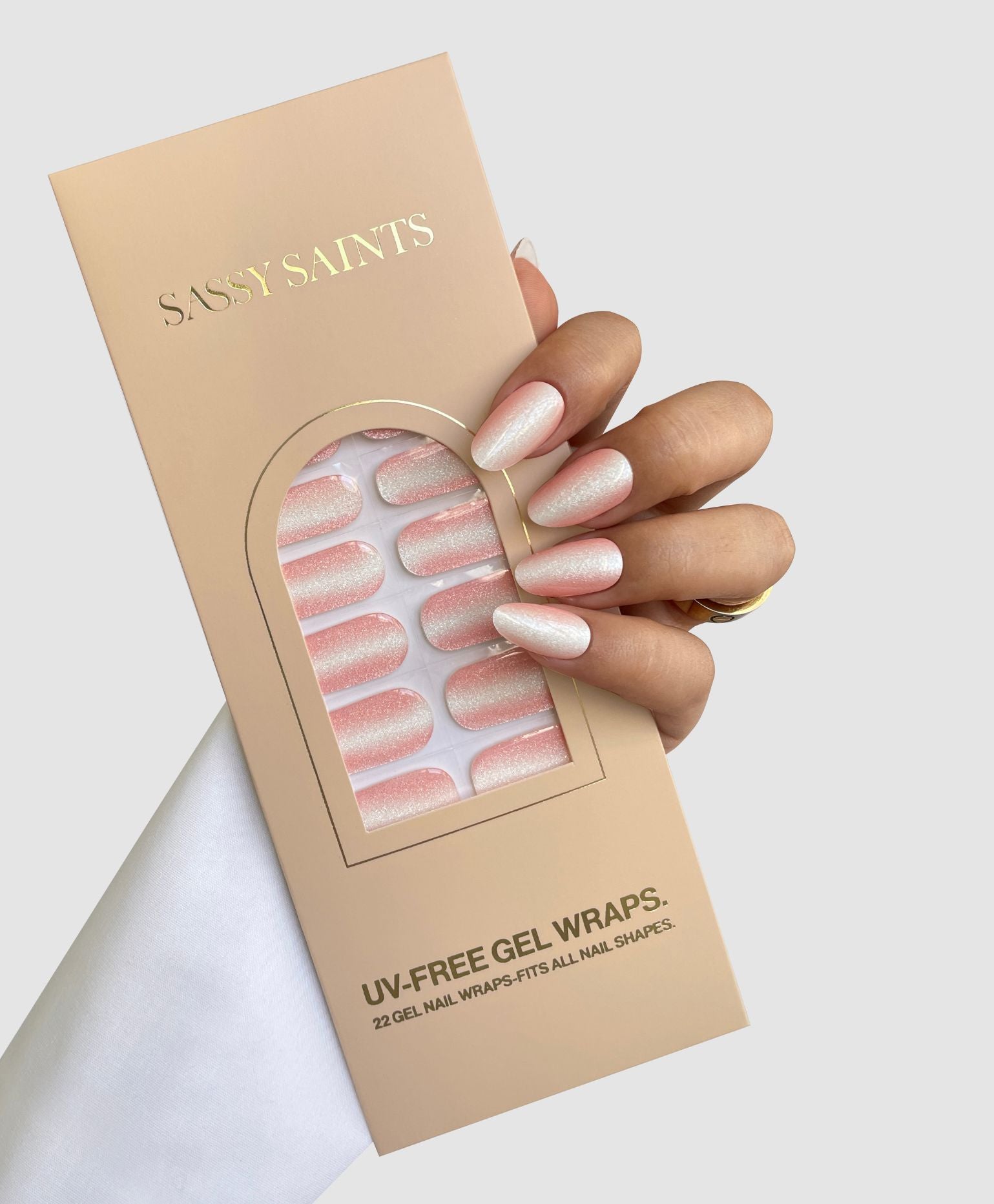 A hand with long, almond-shaped, glittery pink nails holds a beige box labeled “Sassy Saints - Brings The Salon Home” containing Silk Finish Gel Wraps. The box displays 22 shimmery UV-Free Gel Wraps inside.