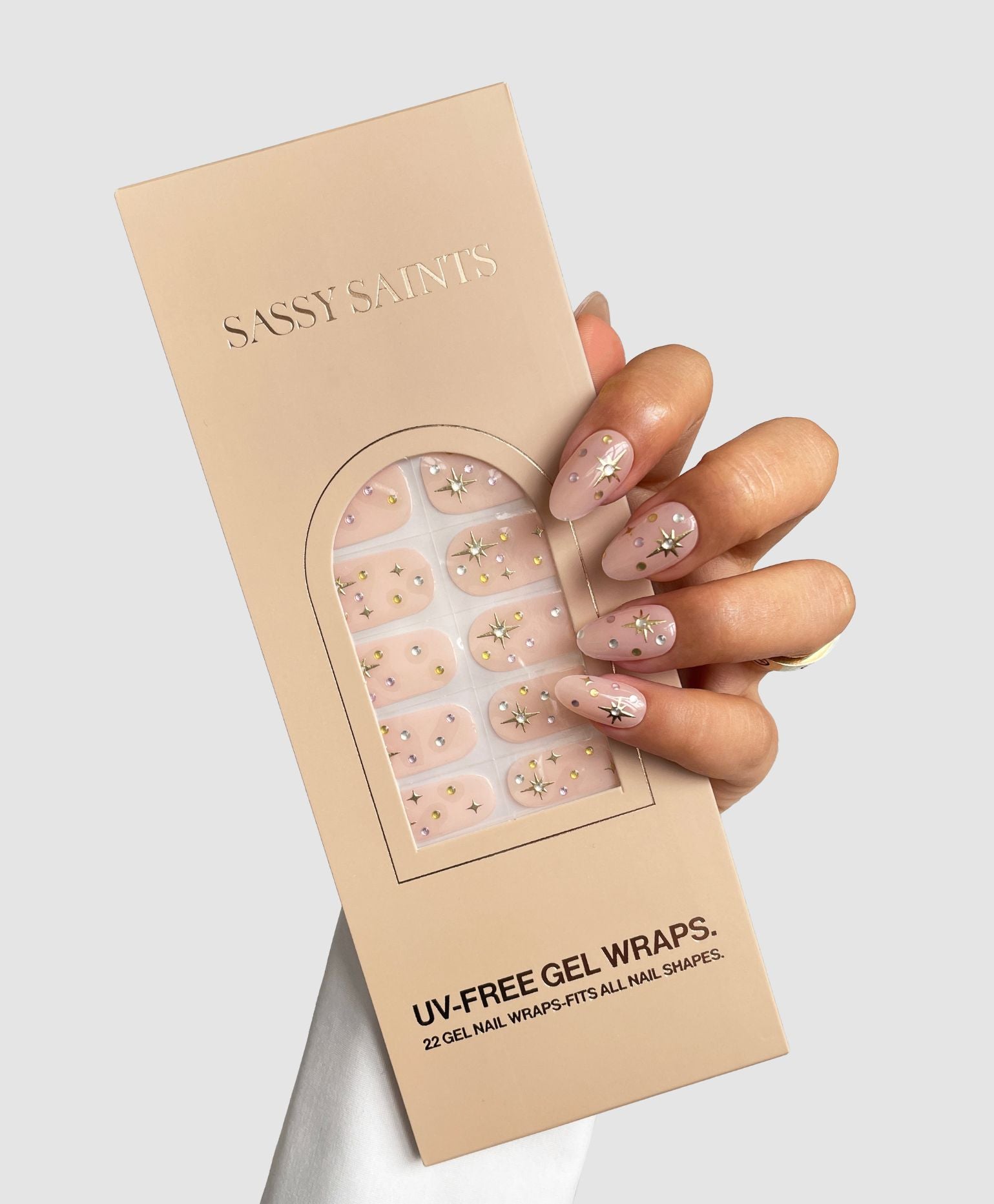 A hand with pink, daisy-patterned nails holds a Sassy Saints - Brings The Salon Home Milkyway Gel Wrap package, displaying matching nail wraps inside with tiny white flowers and gold accents.