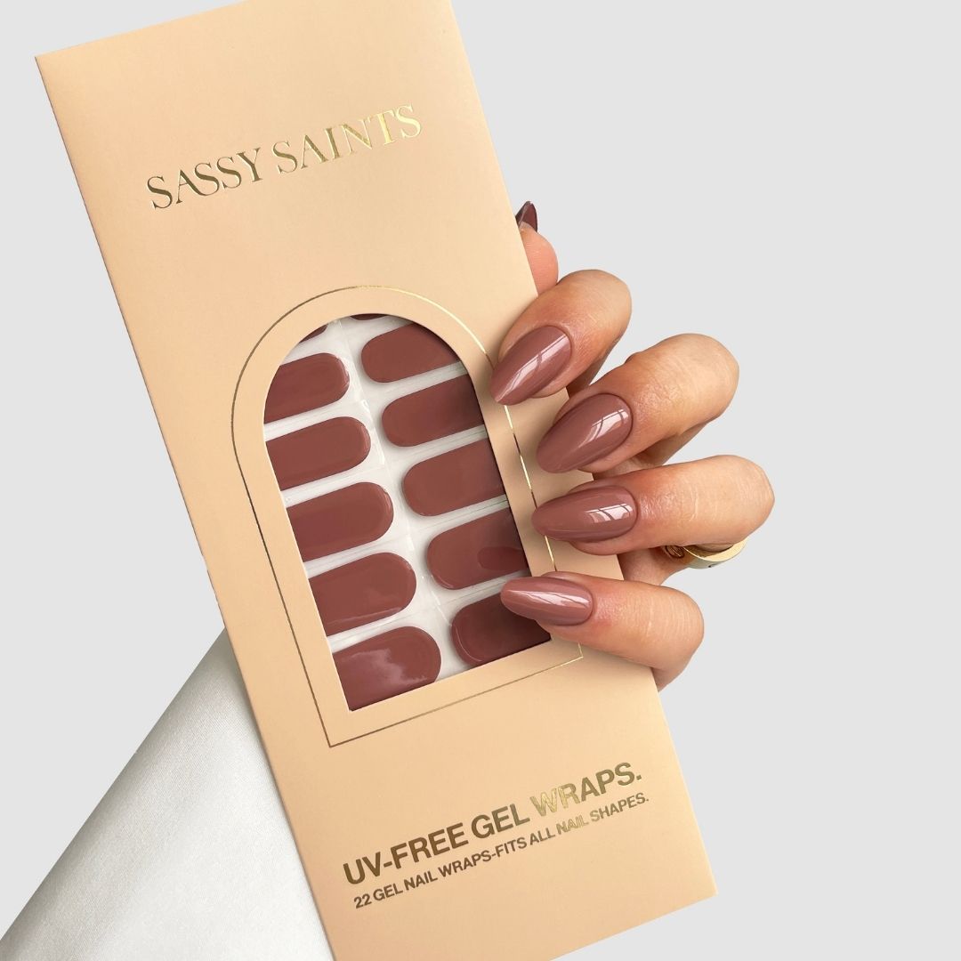 A hand with long, glossy, baked clay-colored nails holds a beige Sassy Saints - Brings The Salon Home package labeled Baked Clay Gel Wrap, containing matching UV-free gel nail wraps. The background is plain light gray.