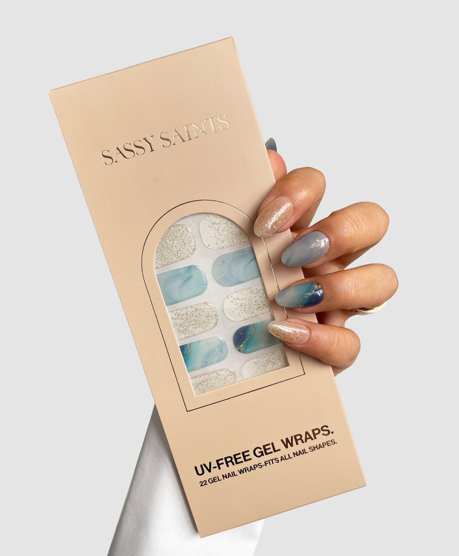 A manicured hand holds a beige Sassy Saints - Brings The Salon Home box containing Ocean Dream Gel Wrap, featuring blue and glittery UV-free gel nail wraps, set against a plain light gray background.