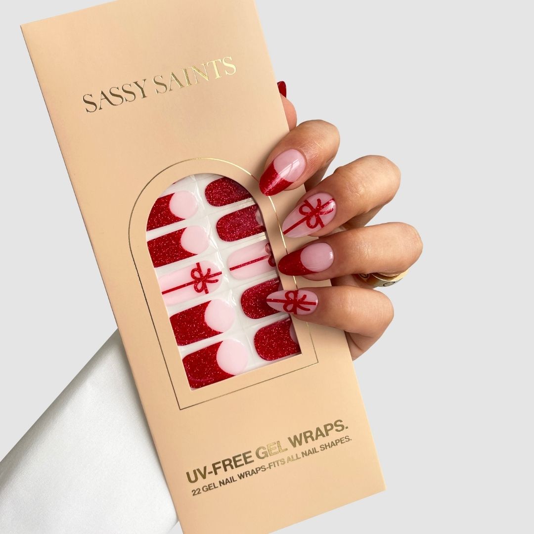 A hand with long, red-glitter and pink nails featuring red bows holds a beige Sassy Saints - Brings The Salon Home Gift Wrapped Gel Wrap box. The box displays the red and pink nail wraps through a clear window.
