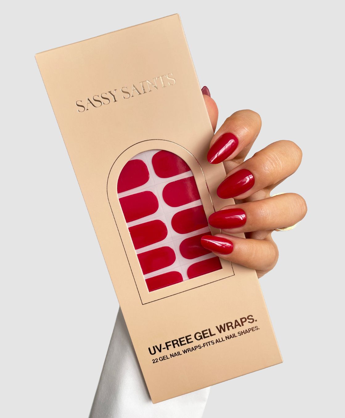 A hand with glossy red nails holds a beige Sassy Saints - Brings The Salon Home package containing Red Alert Gel Wrap UV-free nail wraps, visible through a clear window on the packaging.