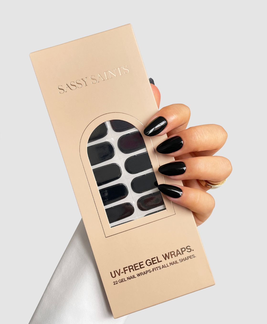 A hand with shiny black polished nails holds a beige box labeled "Liquorice Gel Wrap" from Sassy Saints - Brings The Salon Home, showing black gel nail wraps through a window on the front.