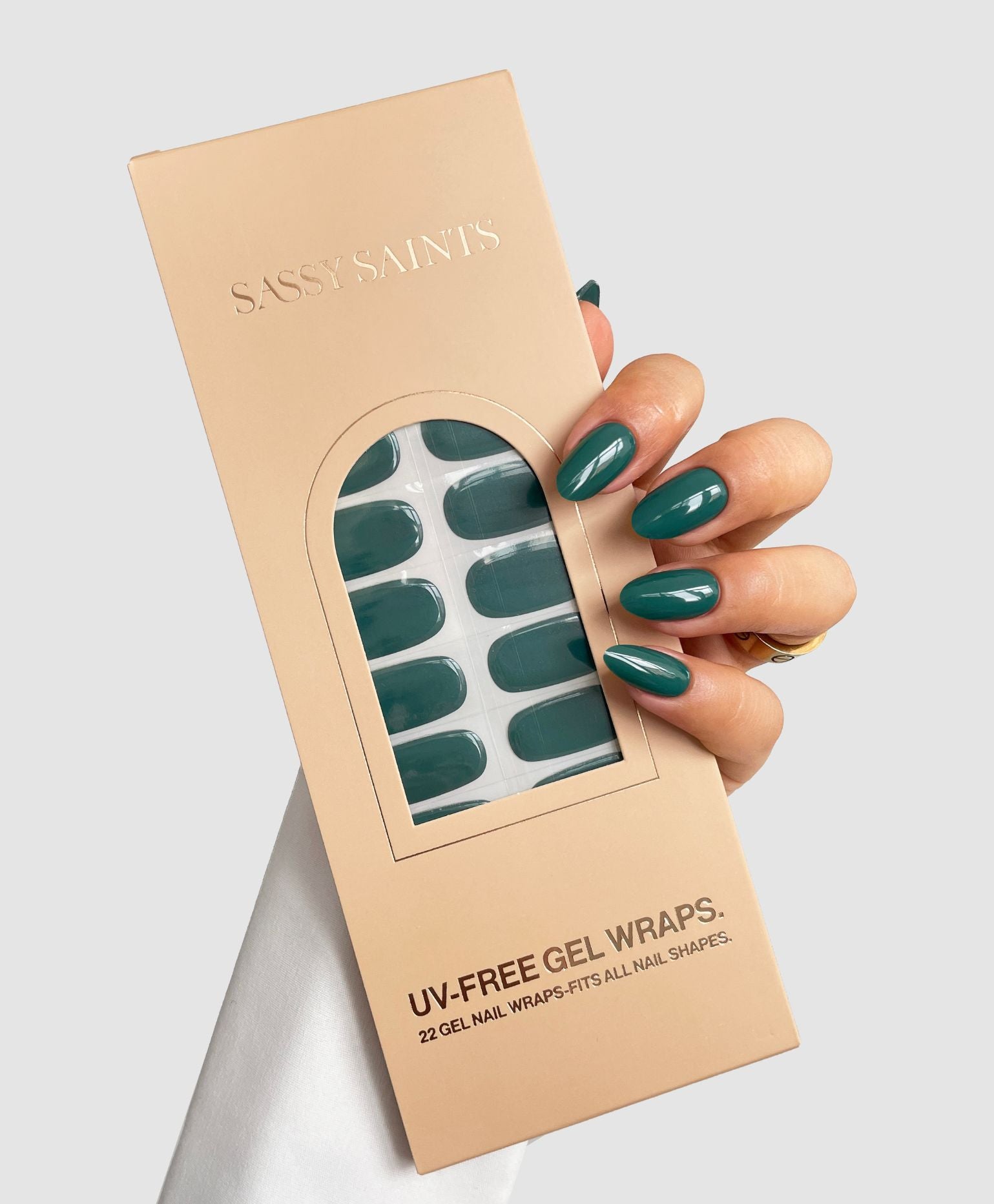 A hand with dark green polished nails holds a beige Sassy Saints – Brings The Salon Home box of Forest Green Gel Wraps, featuring a window that displays the matching UV-free gel nail wraps inside.