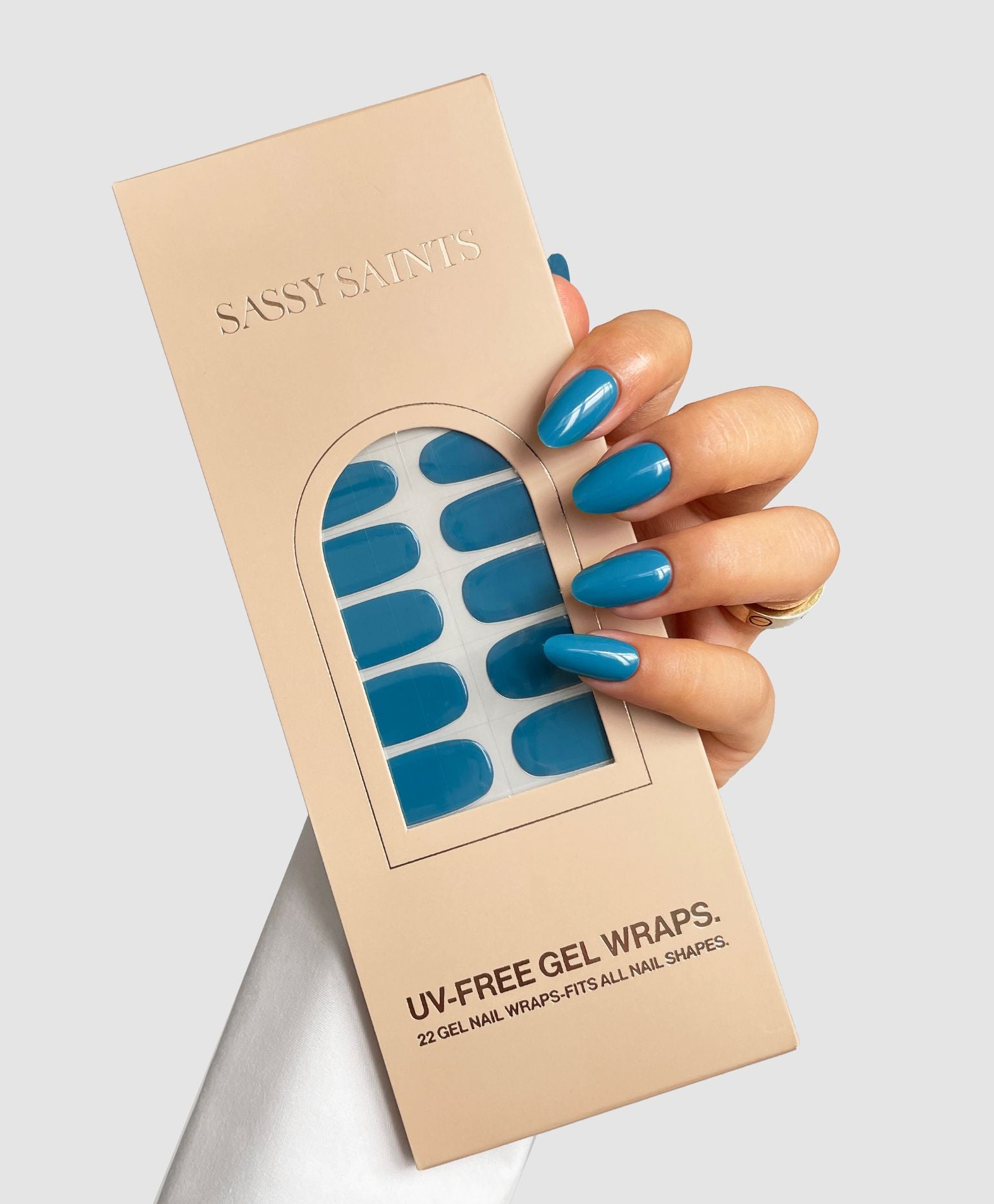 A hand with blue Tonal Drift Gel Wrap nails holds a beige Sassy Saints - Brings The Salon Home box featuring a window that displays the matching blue UV-free gel nail wraps inside.