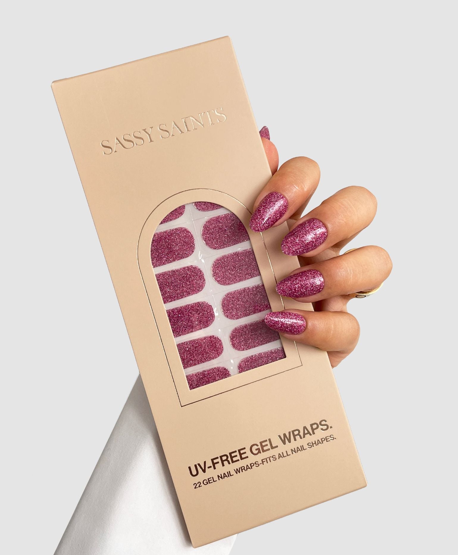 A hand with sparkly pink gel nails holds a beige box labeled Doll Moment Gel Wrap by Sassy Saints - Brings The Salon Home, revealing glittery pink nail wraps inside.
