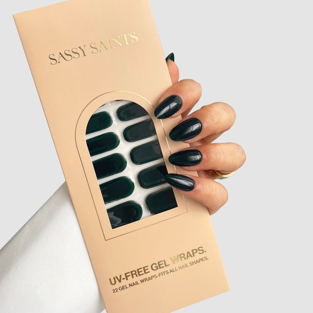 A hand with long, glossy black nails holds a pink “Sassy Saints - Brings The Salon Home” box labeled “Velvet Fir (Dark Green),” featuring a window that displays the dark green gel nail wraps inside.