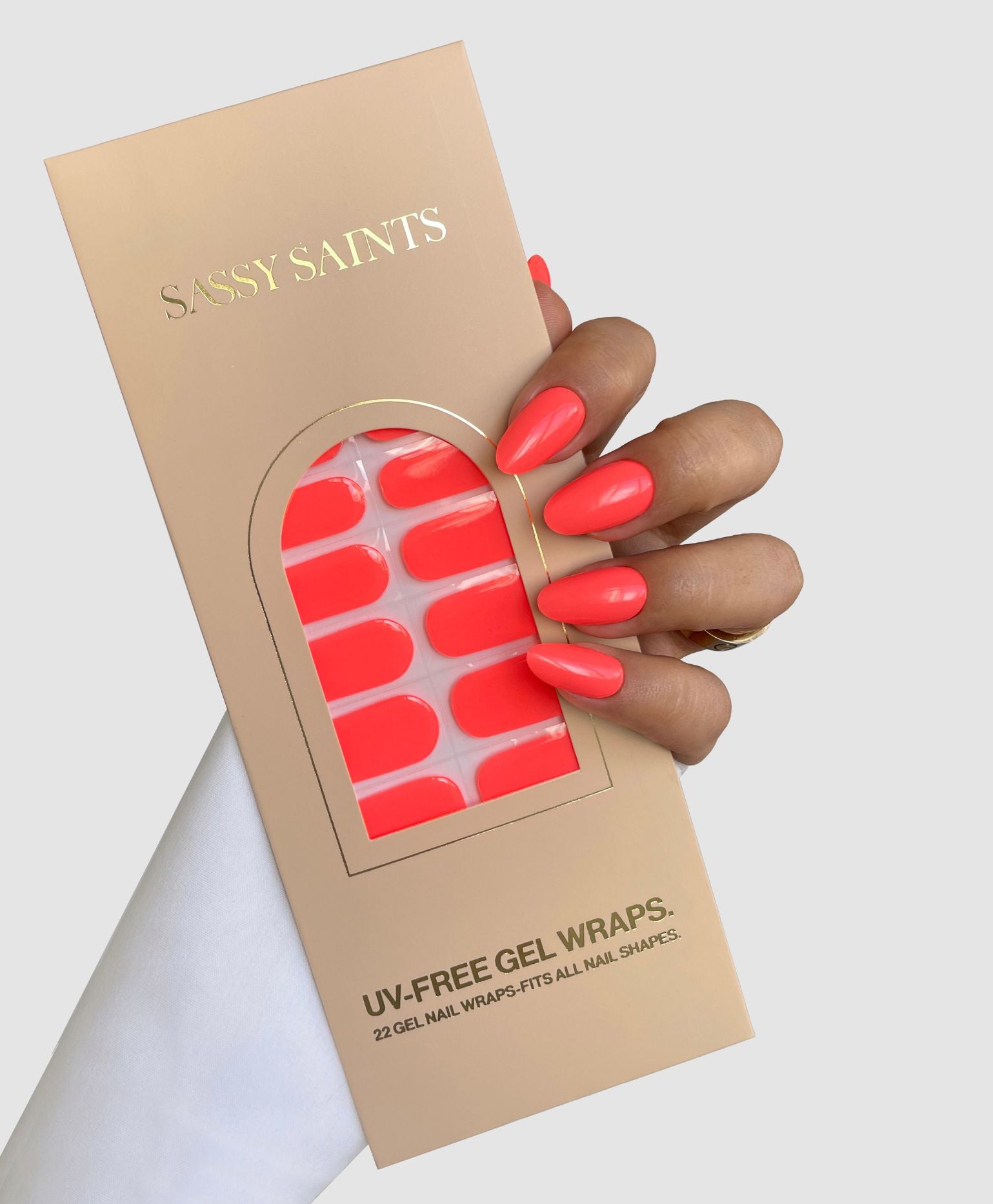 A hand with bright coral nails holds a beige Sassy Saints - Brings The Salon Home package labeled Coral Kiss Gel Wrap, featuring a clear window that displays the matching UV-free gel nail wraps inside.