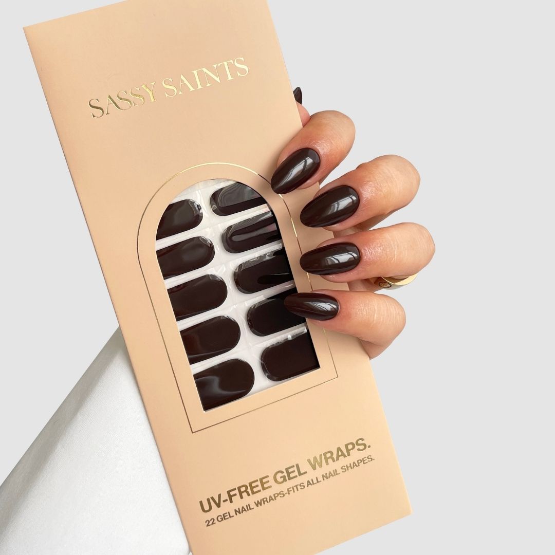 A hand with almond-shaped, dark brown nails holds a beige package of Sassy Saints – Brings The Salon Home Cocoa Luxe (Dark Brown) UV-free gel nail wraps, with a window displaying the matching wraps inside.