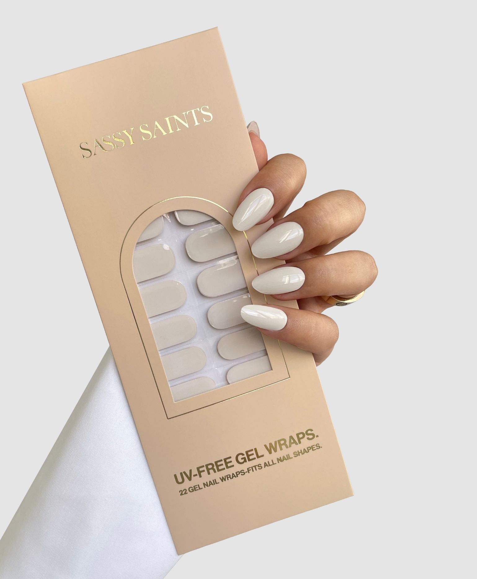 A hand with glossy, pale nude Blush Milk Gel Wraps by Sassy Saints - Brings The Salon Home holds a beige package containing 22 matching UV-free gel nail wraps. The background is plain light gray.