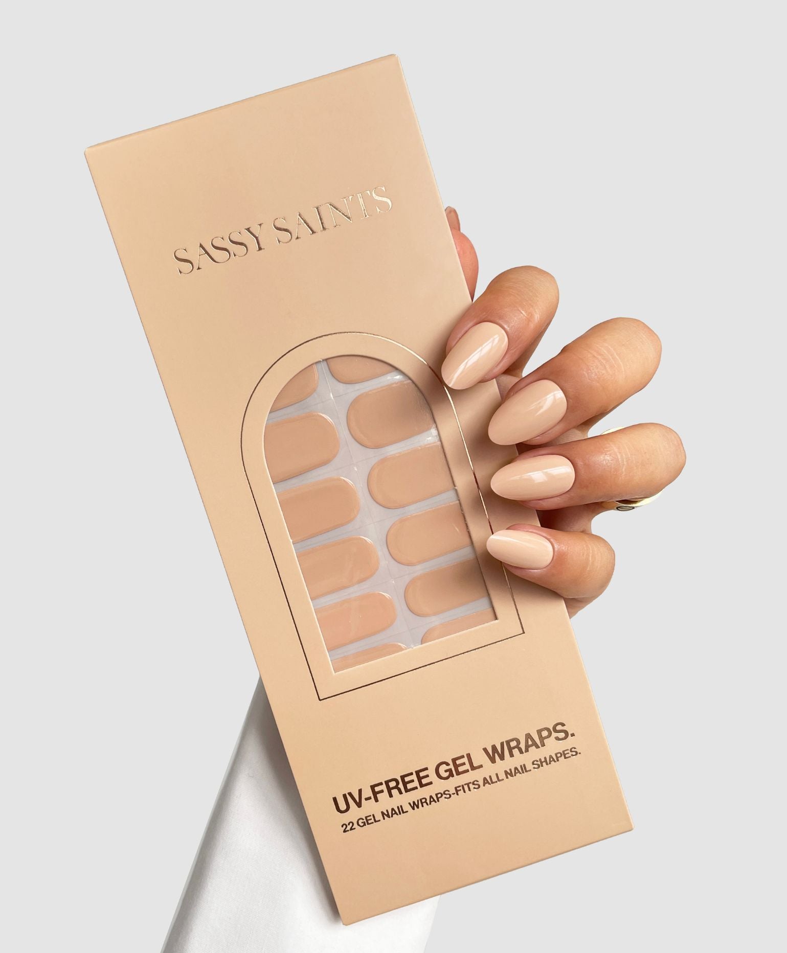 A hand with nude Naked Ambition Gel Wraps holds a beige Sassy Saints - Brings The Salon Home box labeled UV-Free Gel Wraps containing matching wraps, set against a light gray background.