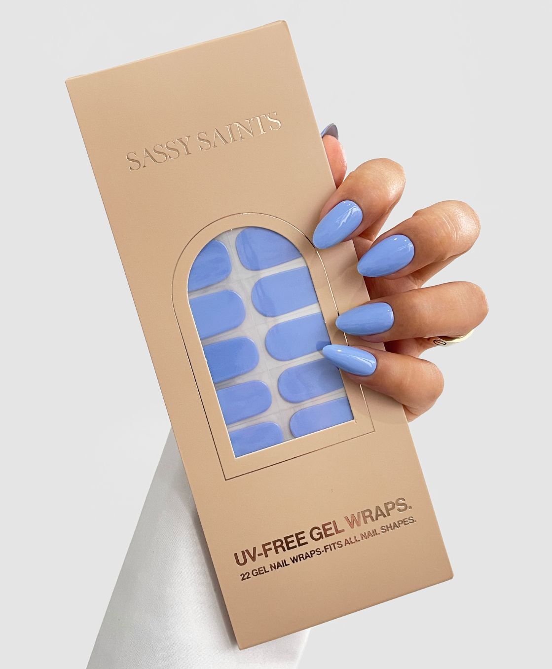 A hand with long, glossy, light blue nails holds a beige package of Sassy Saints - Brings The Salon Home Iris Bleu Gel Wraps. The pack contains 22 UV-free gel nail wraps in light blue and features a window showing the wraps inside.
