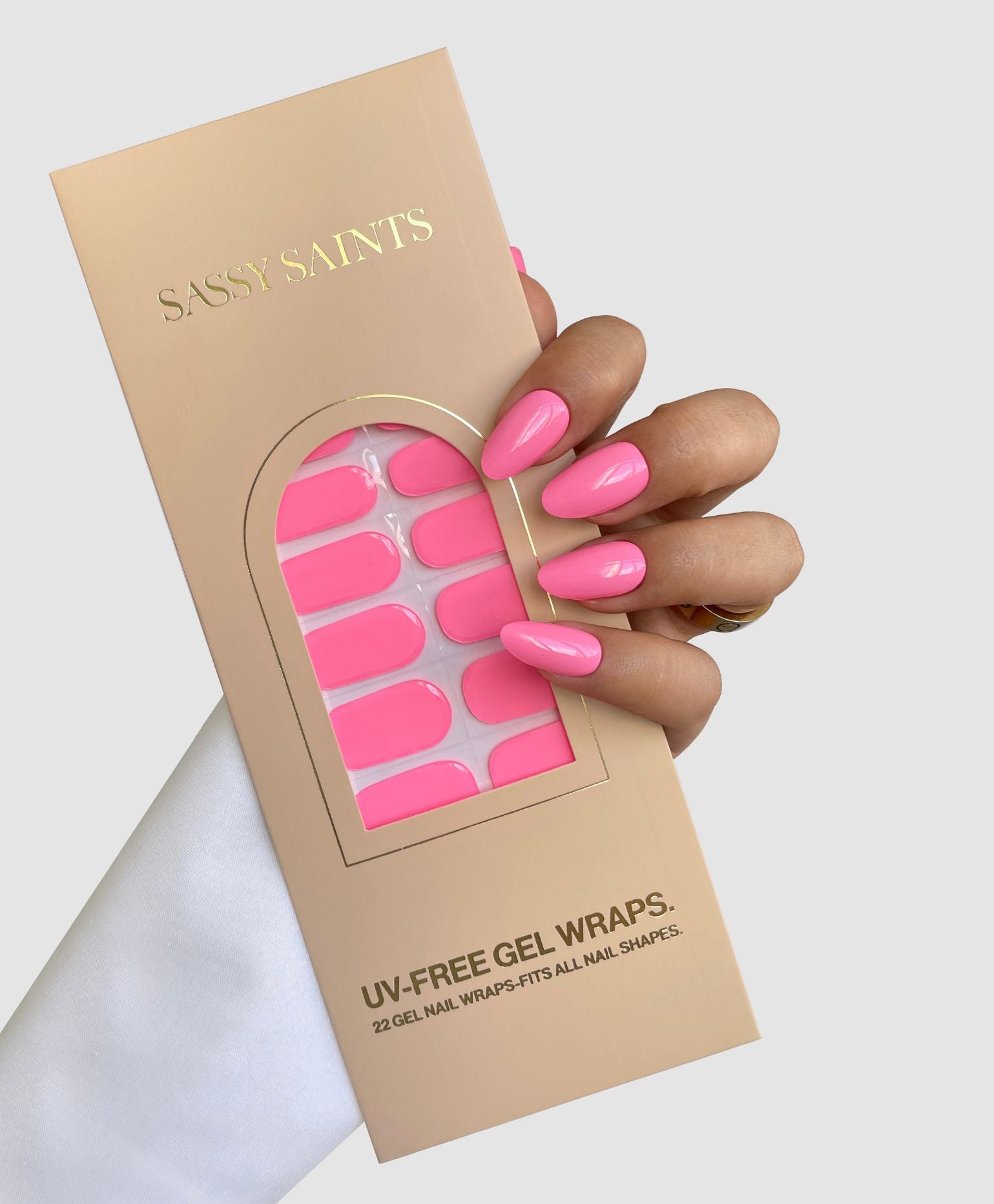 A hand with long, glossy, bright pink nails holds a beige box of Sassy Saints - Brings The Salon Home Sweet Beat Gel Wraps. The box features gold lettering and a clear window displaying the matching pink nail wraps.