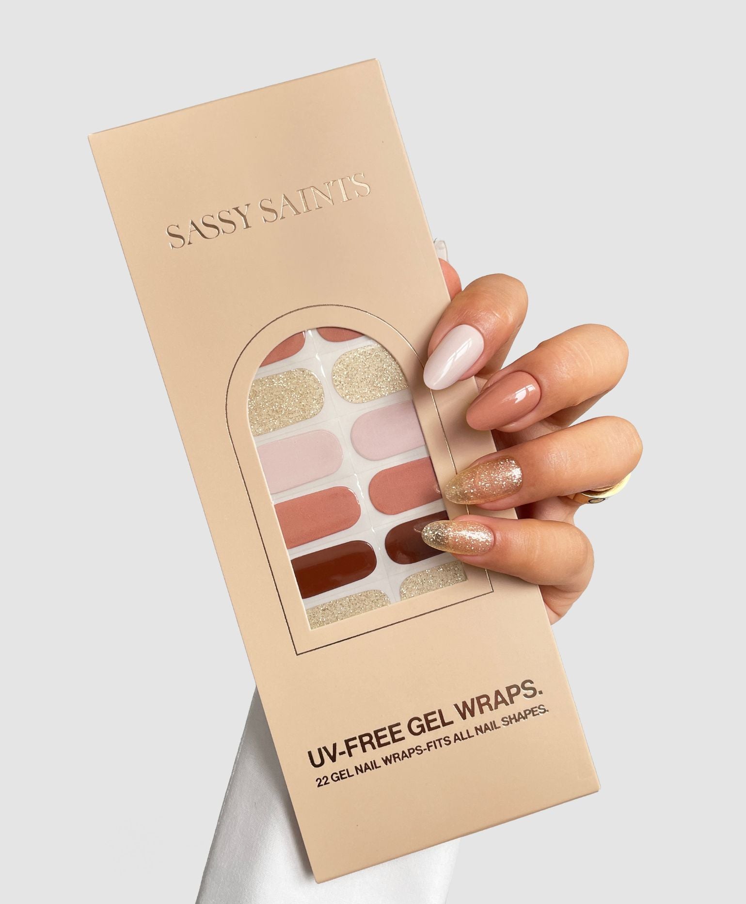 A hand with manicured nails holds a beige box of Sassy Saints - Brings The Salon Home Bare Minimum Gel Wraps, displaying pink, nude, and glitter designs through a clear window on the package.