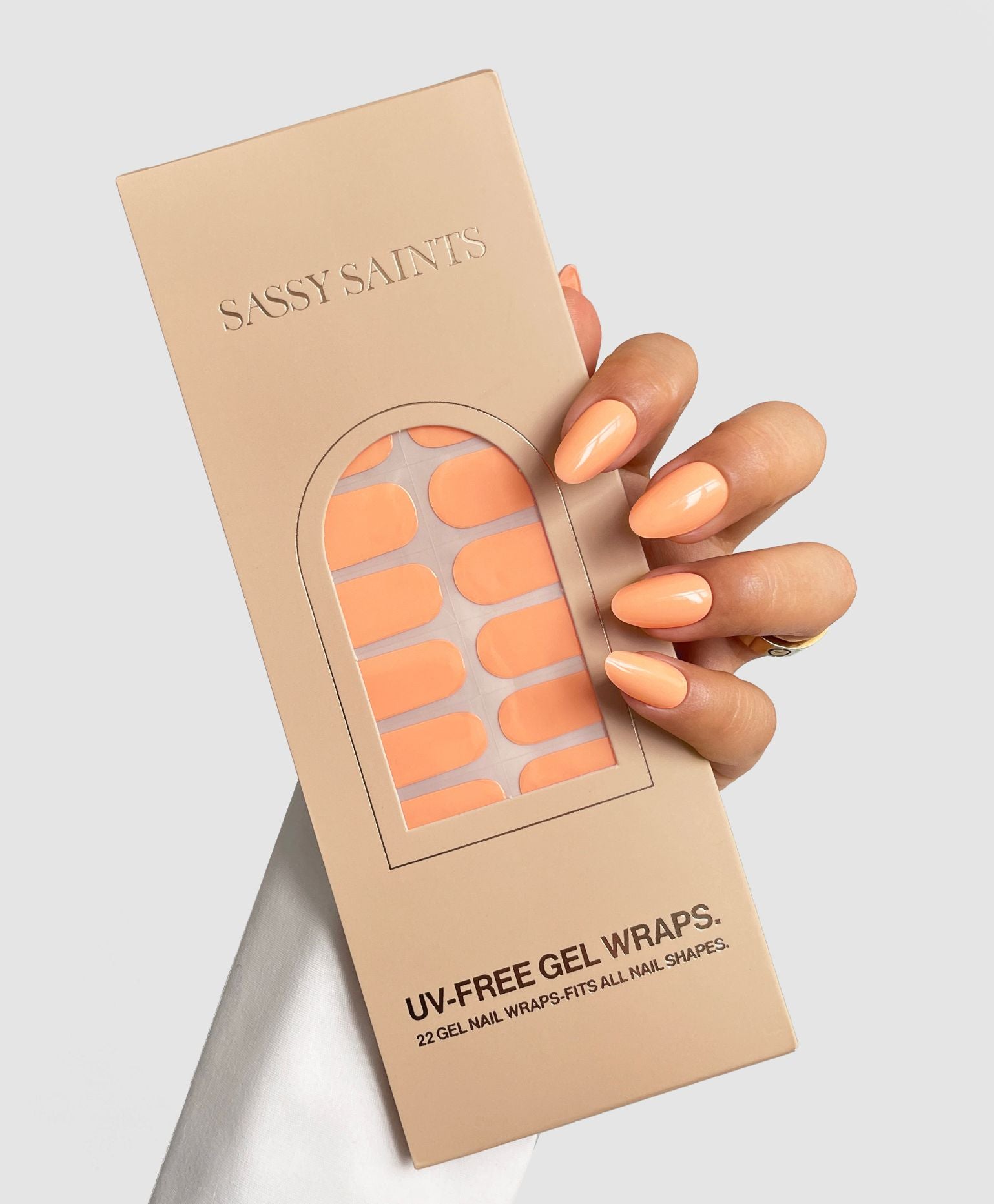 A hand with peach-colored, manicured nails holds a box of Sassy Saints - Brings The Salon Home Peach Glow Gel Wraps in a matching shade against a light gray background.