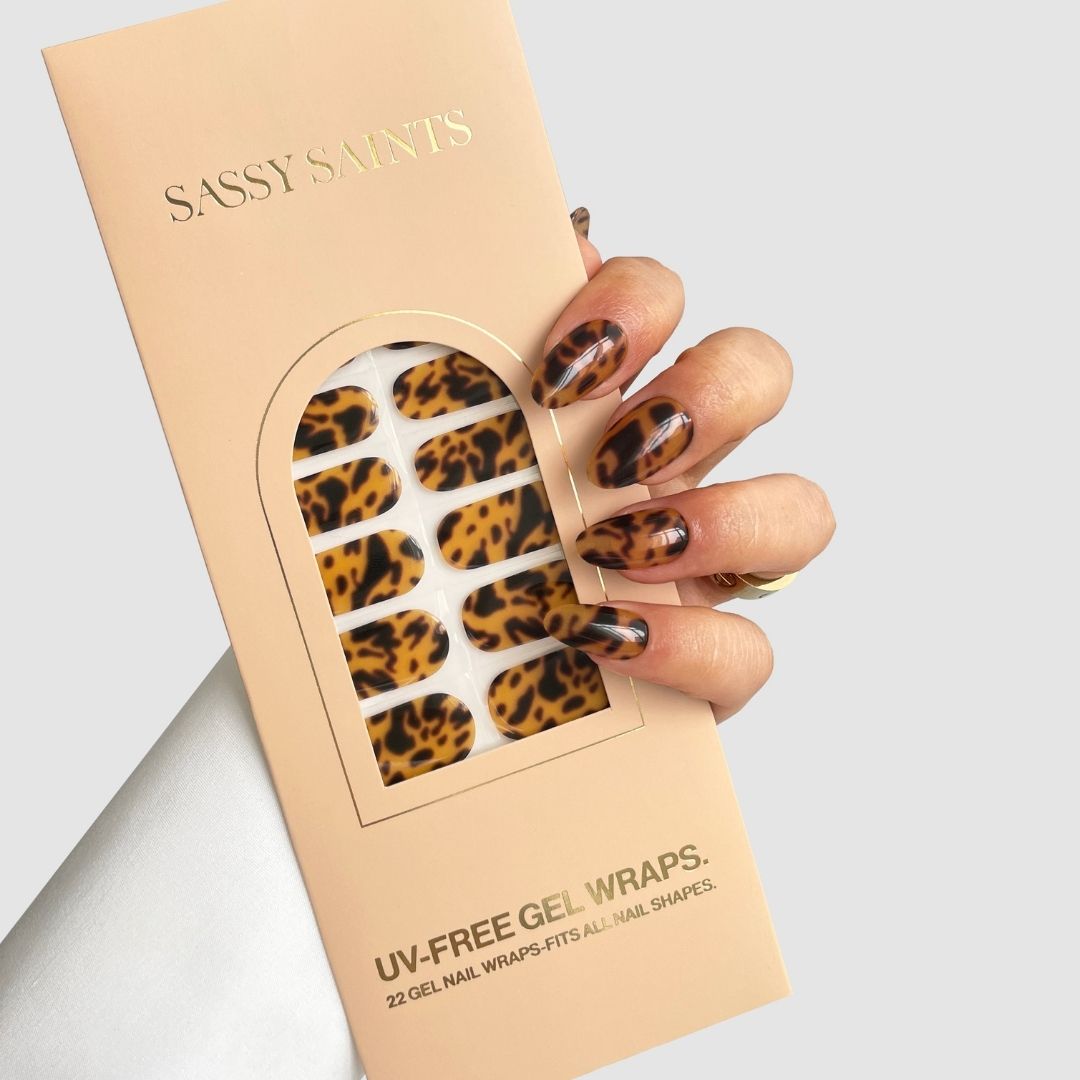 A hand with brown and gold leopard-print nails holds a Sassy Saints - Brings The Salon Home Shell Luxe Gel Wrap package displaying 22 animal-print, UV-free gel nail wraps on a neutral background.
