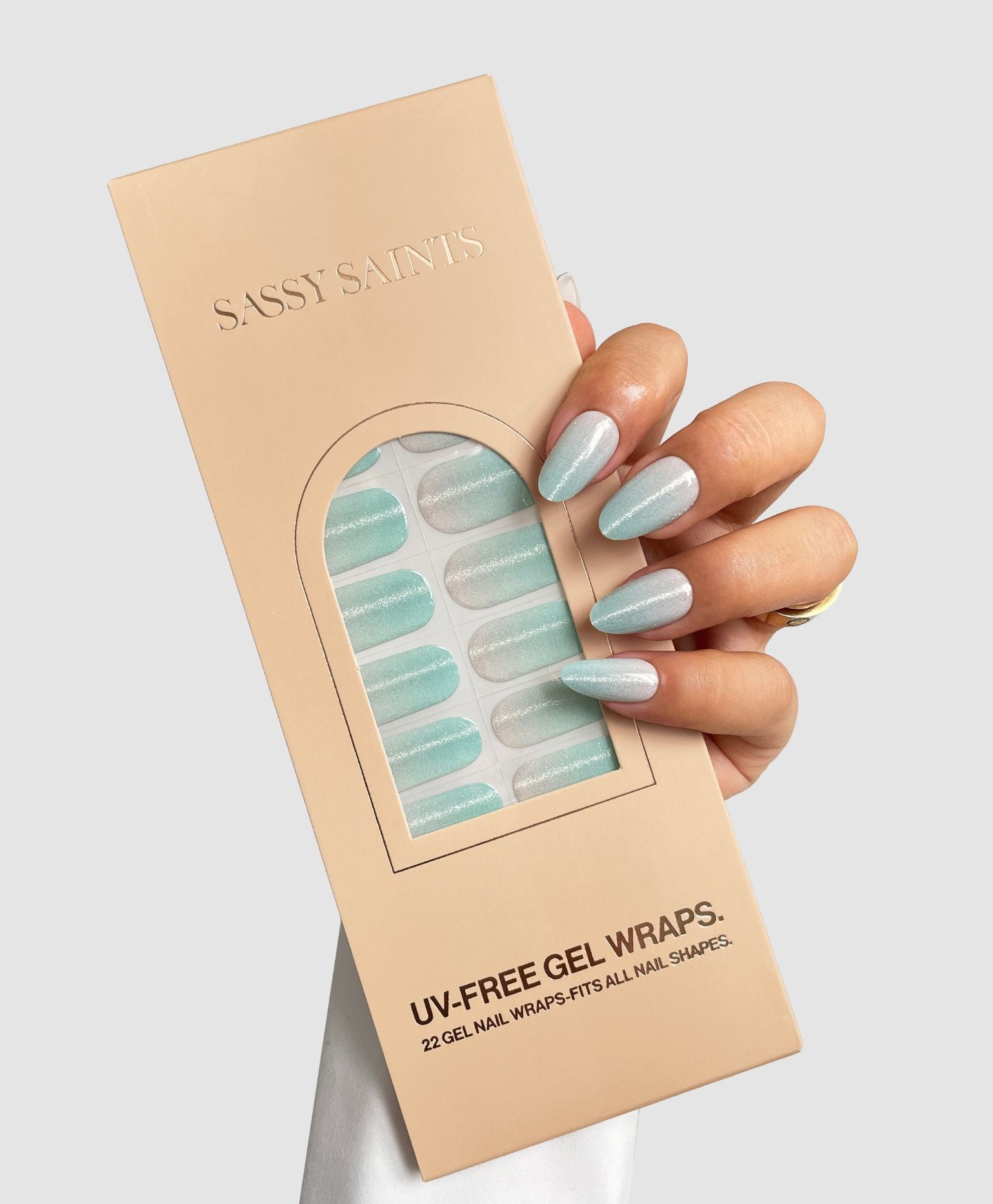 A hand with light blue and silver gradient nails holds a beige Sassy Saints - Brings The Salon Home package labeled Frozen Glow Gel Wrap, featuring UV-free gel nail wraps in matching pastel blue and silver shades.
