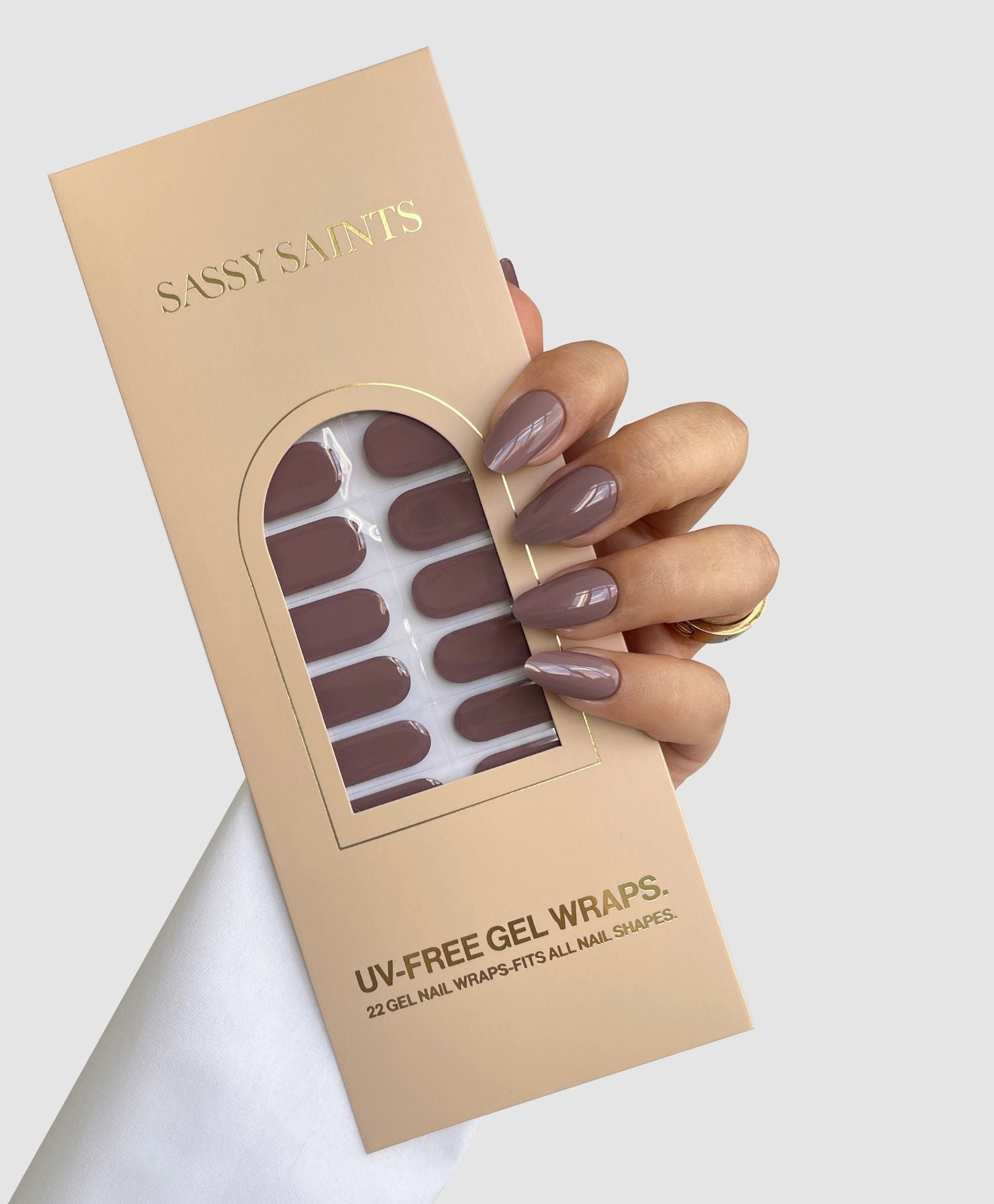 A hand with glossy mauve gel nails holds a Sassy Saints - Brings The Salon Home Dusky Mauve Gel Wrap box, showing matching nail wraps in a clear window on the front.