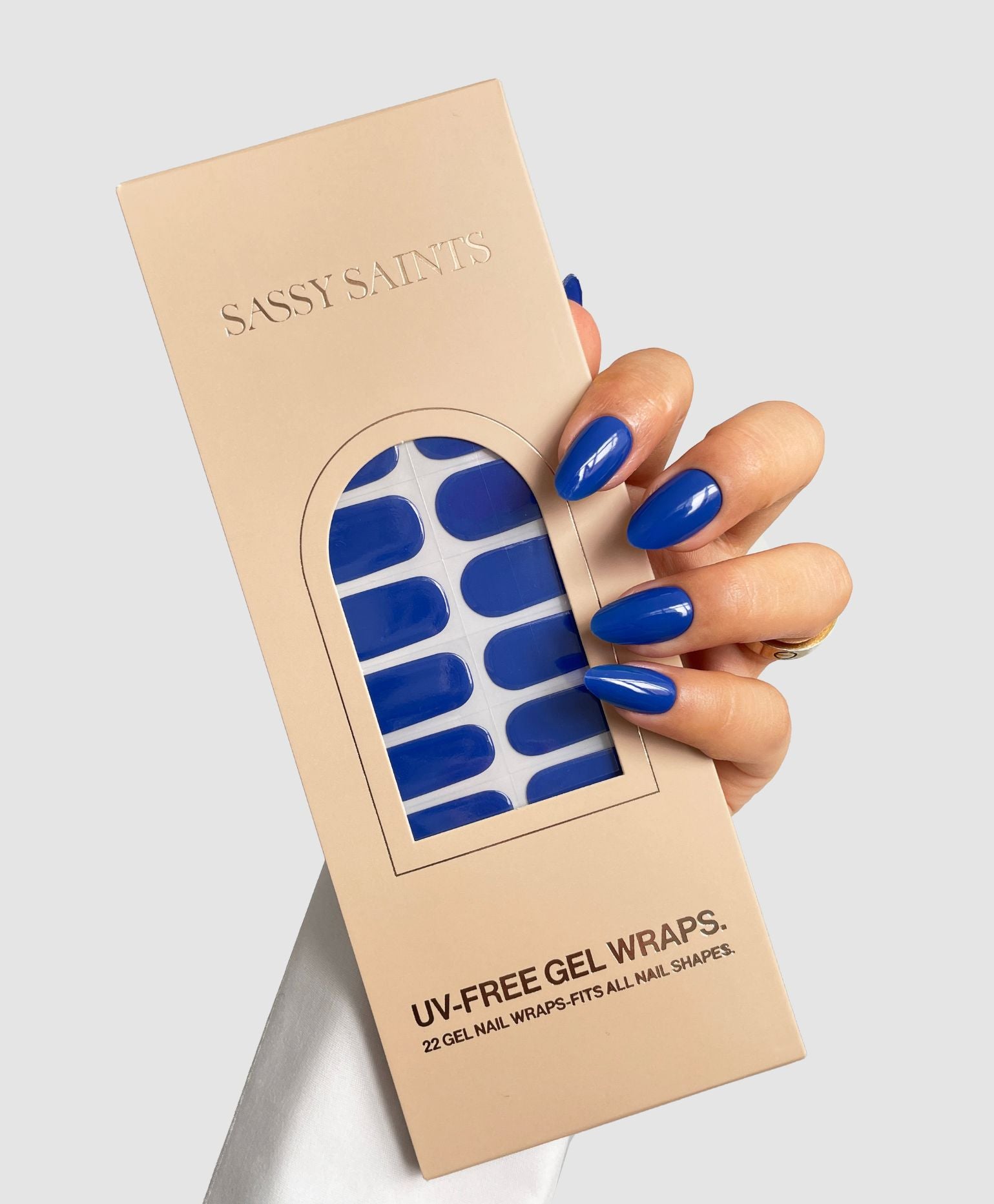 A hand with long, glossy blue nails holds a beige box labeled "Blue Me Away Gel Wrap" by Sassy Saints – Brings The Salon Home, showing matching blue gel nail wraps inside.