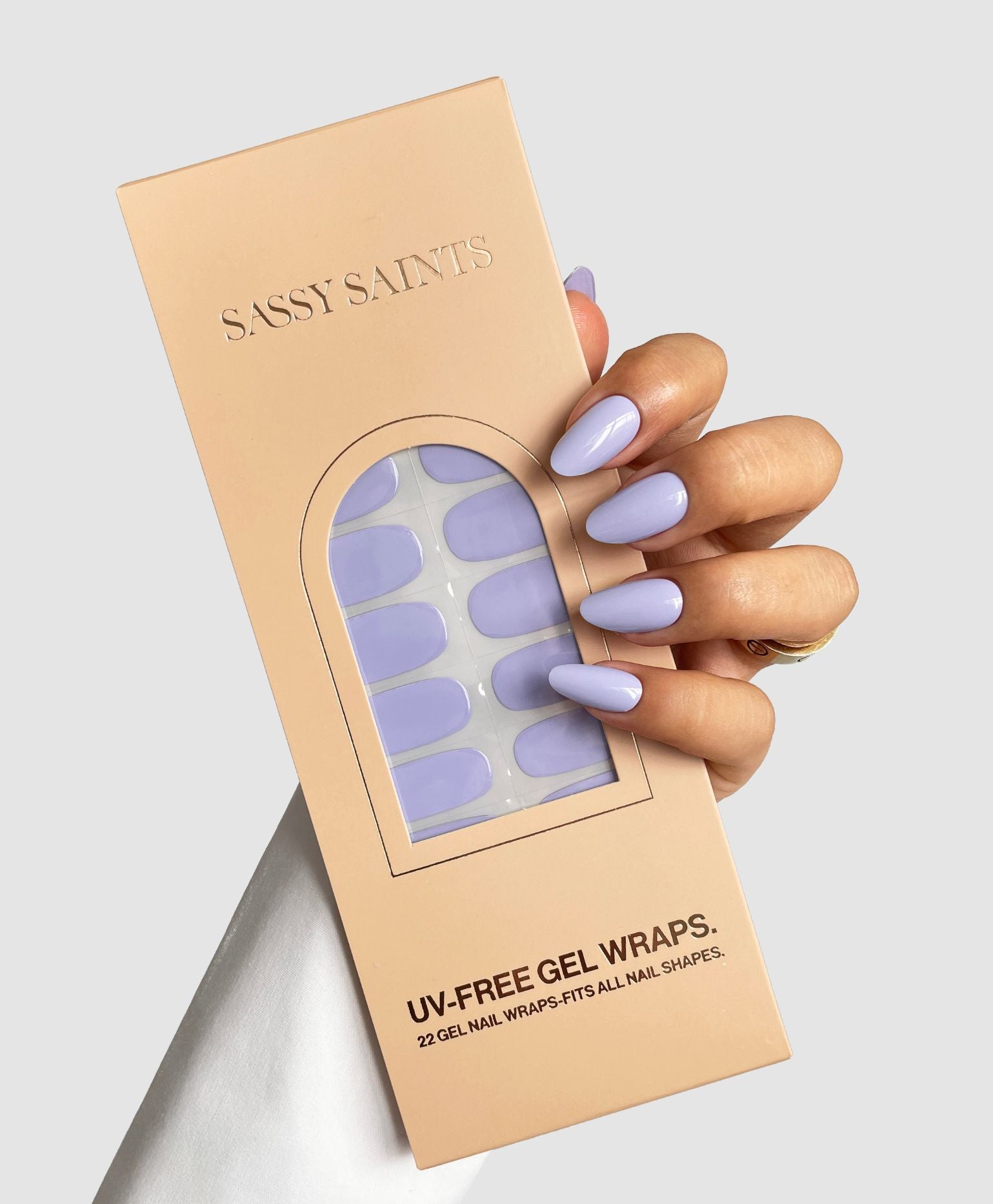 A hand with long, glossy lavender nails holds a beige Sassy Saints – Brings The Salon Home box labeled "Lavender Blush Gel Wrap" with a clear window showing the matching UV-free gel nail wraps inside.