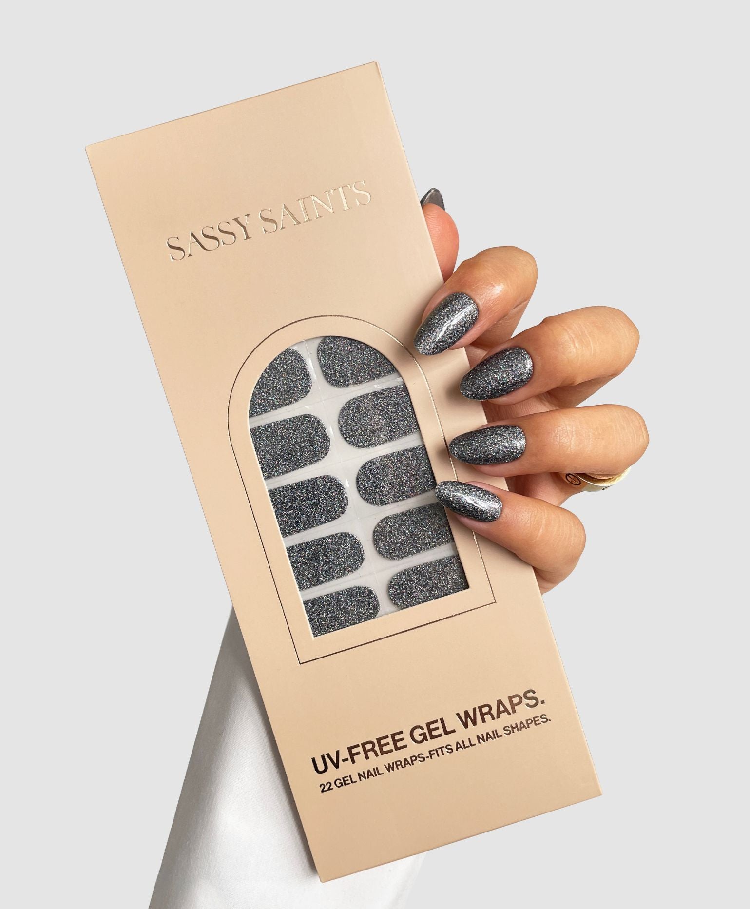 A hand with sparkly silver Midnight Glimmer Gel Wraps holds a beige Sassy Saints - Brings The Salon Home package, showing dark glittery nail strips inside.