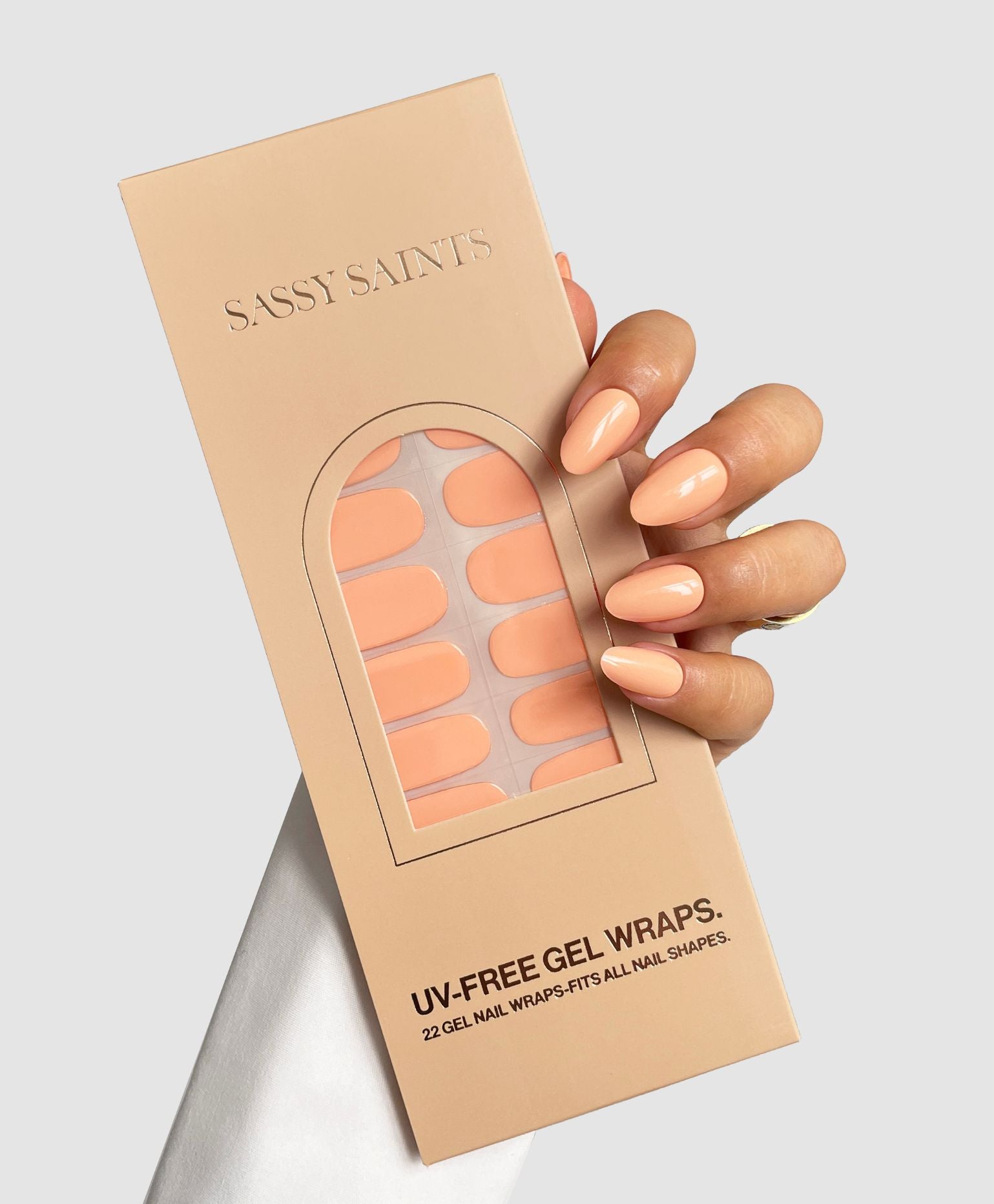 A hand with glossy apricot nails holds a box of Sassy Saints - Brings The Salon Home Apricot Mood Gel Wraps. The box contains 22 UV-free wraps for all nail shapes. Background is light gray.