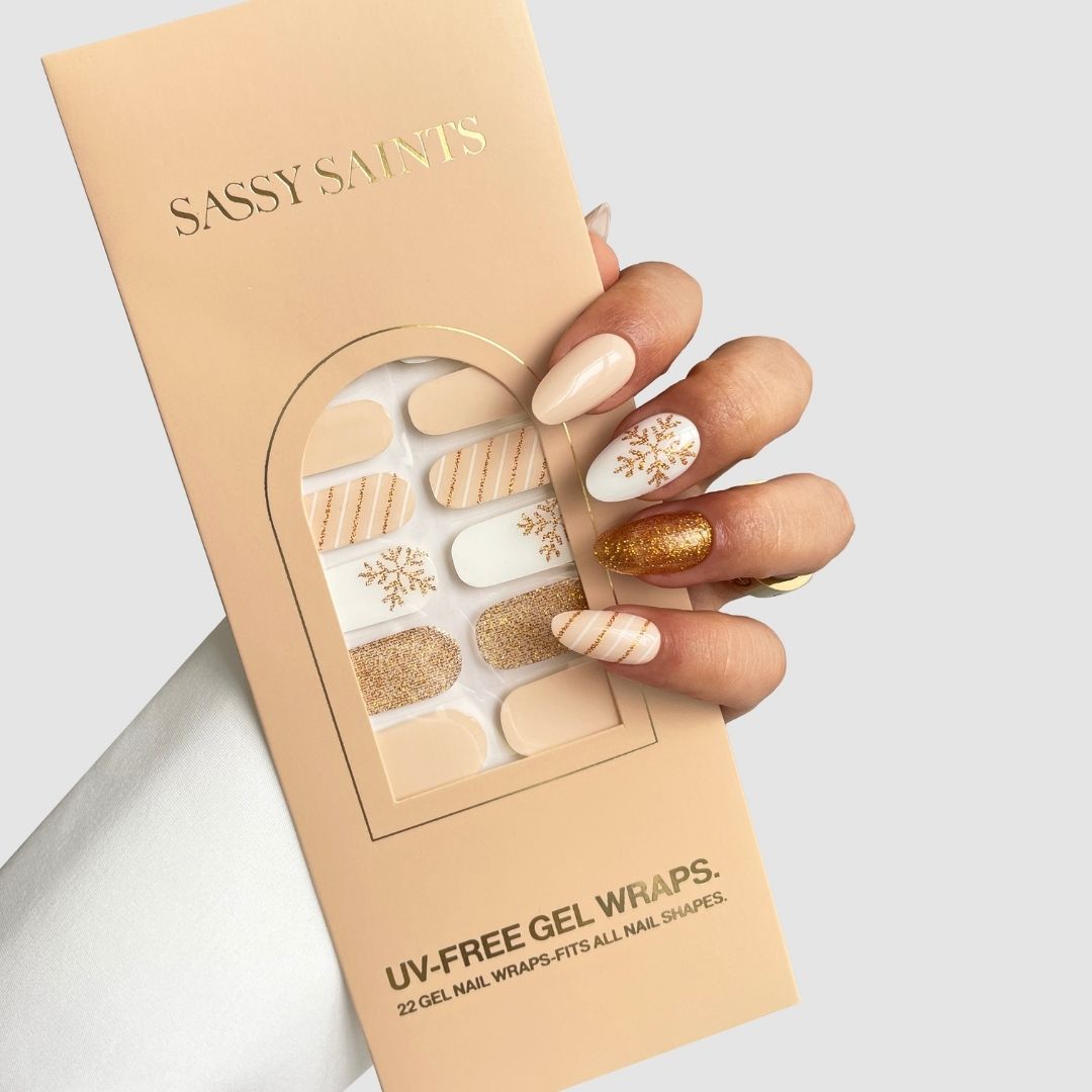 A manicured hand showcasing neutral, gold, and glittery nail art holds a Sassy Saints - Brings The Salon Home Golden Flurry Gel Wrap package with beige, gold, and snowflake designs against a light gray background.
