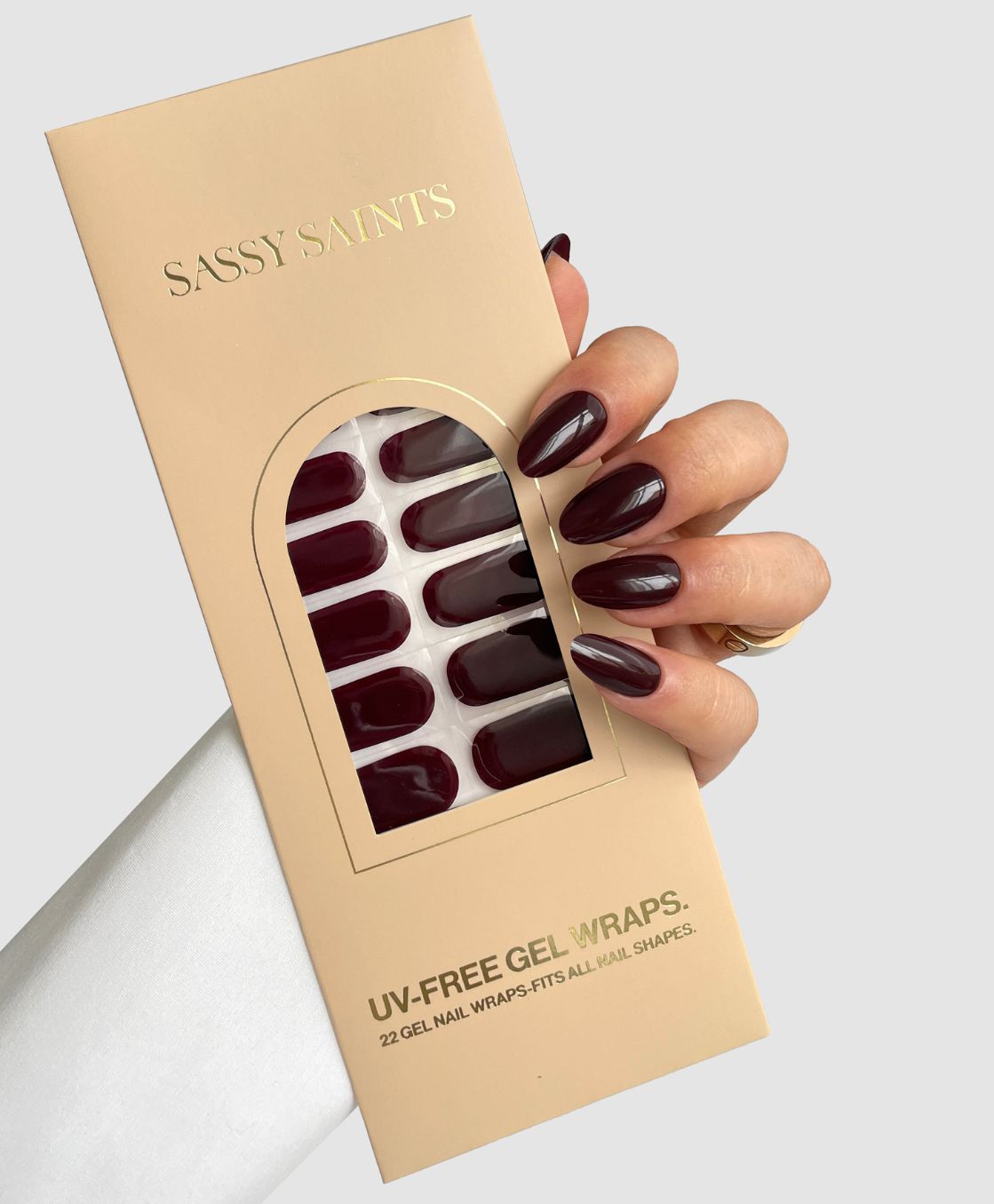 A hand with glossy dark red "Dark Current" nails holds a beige Sassy Saints – Brings The Salon Home UV-Free Gel Wraps package, showing dark gel wraps through a clear window, set against a light gray background.