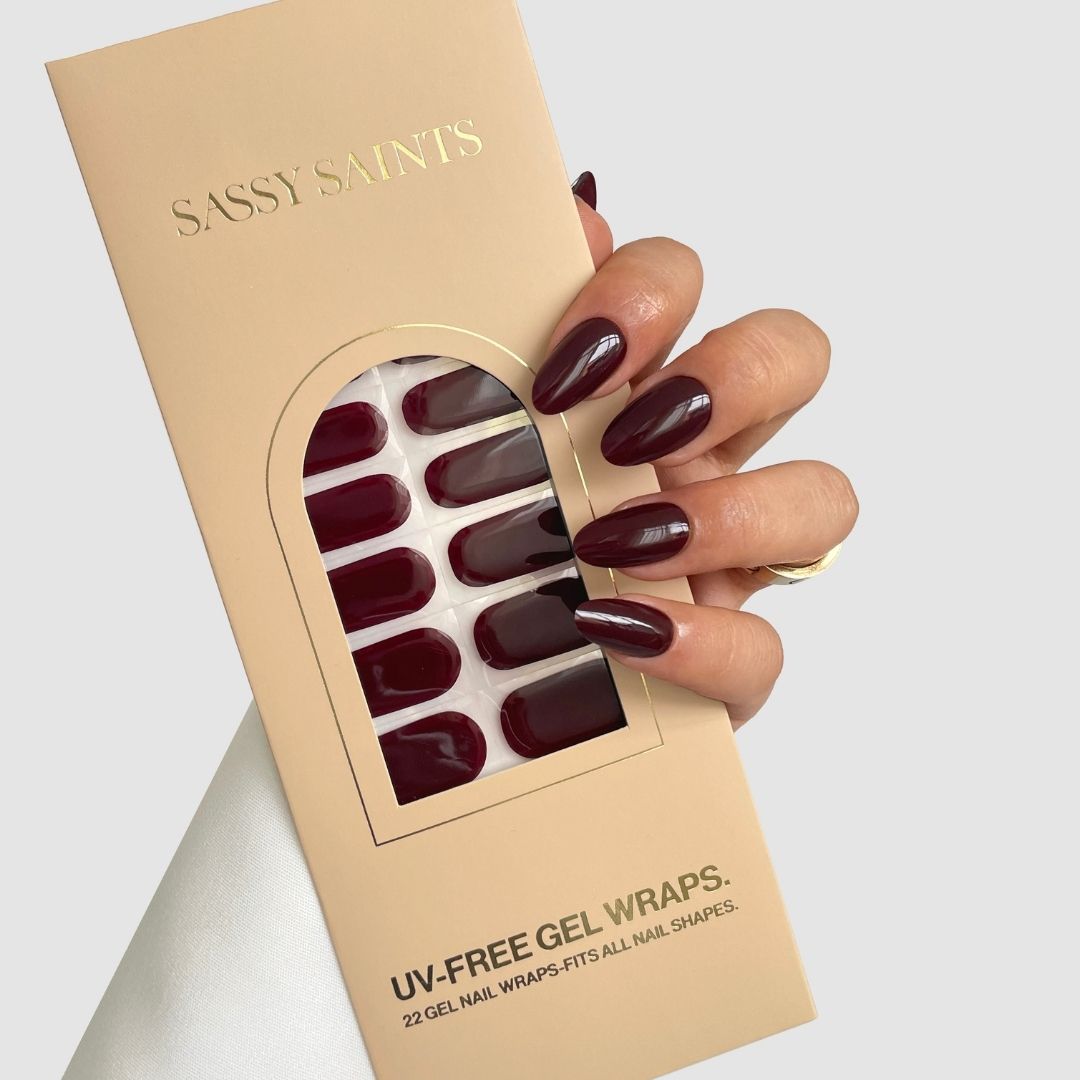 A hand with long, dark red nails holds a beige Sassy Saints – Brings The Salon Home box labeled "Dark Current," displaying matching UV-free gel nail wraps through a clear window.