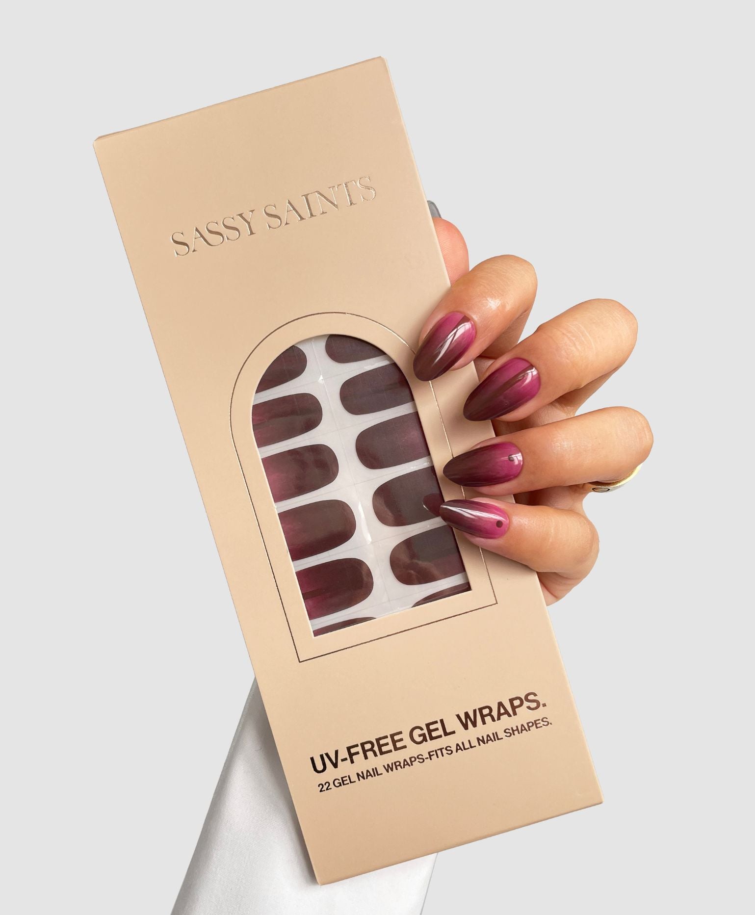 A hand with glossy, mauve nails holds a beige Sassy Saints - Brings The Salon Home box of Cherry Smoke Gel Wraps. The UV-free gel wraps, visible through a clear window, match the nail color.