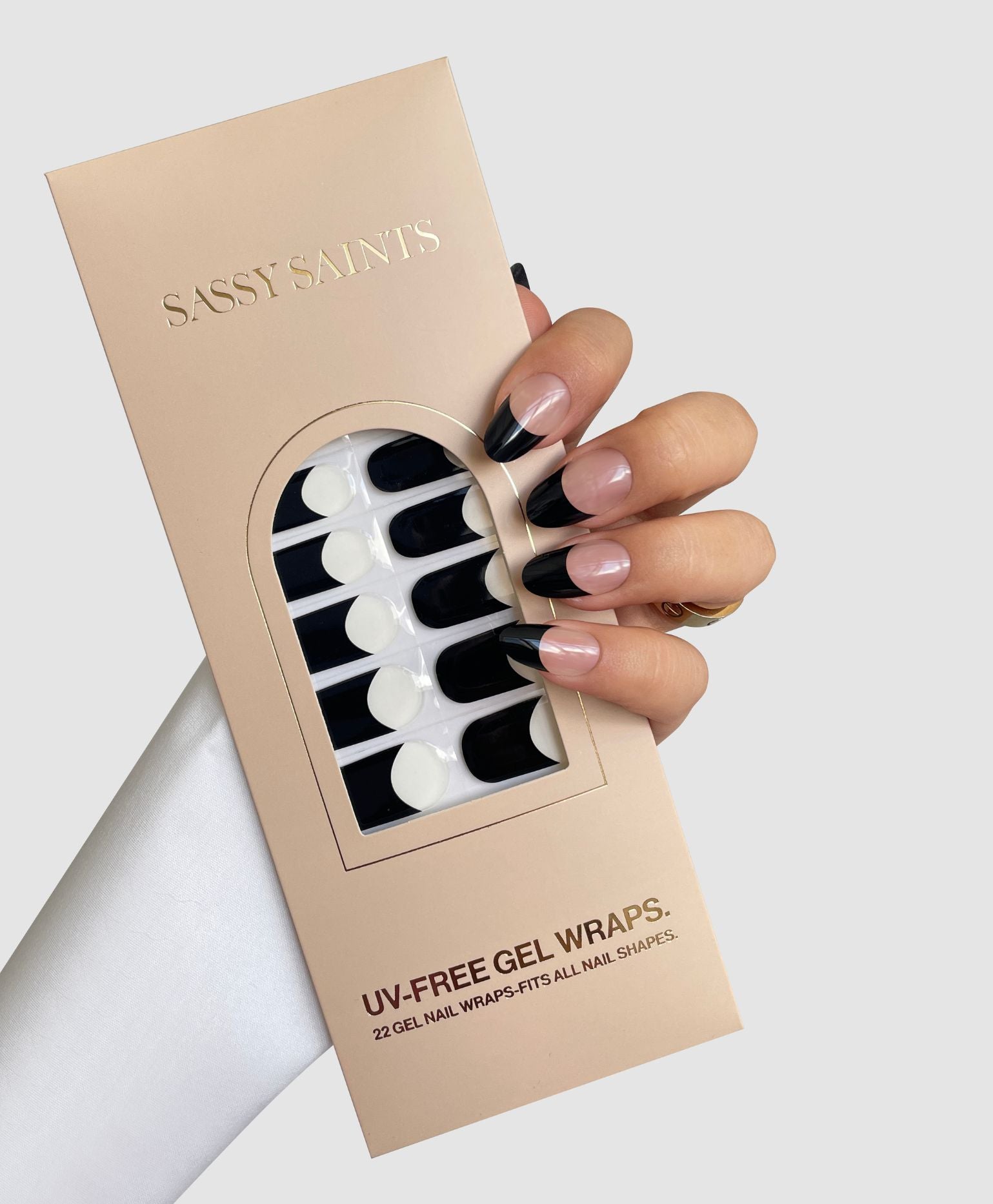 A hand with black and nude French tip nails holds a box of Sassy Saints - Brings The Salon Home Midnight French Gel Wraps, revealing black and white gel wraps inside beige packaging.