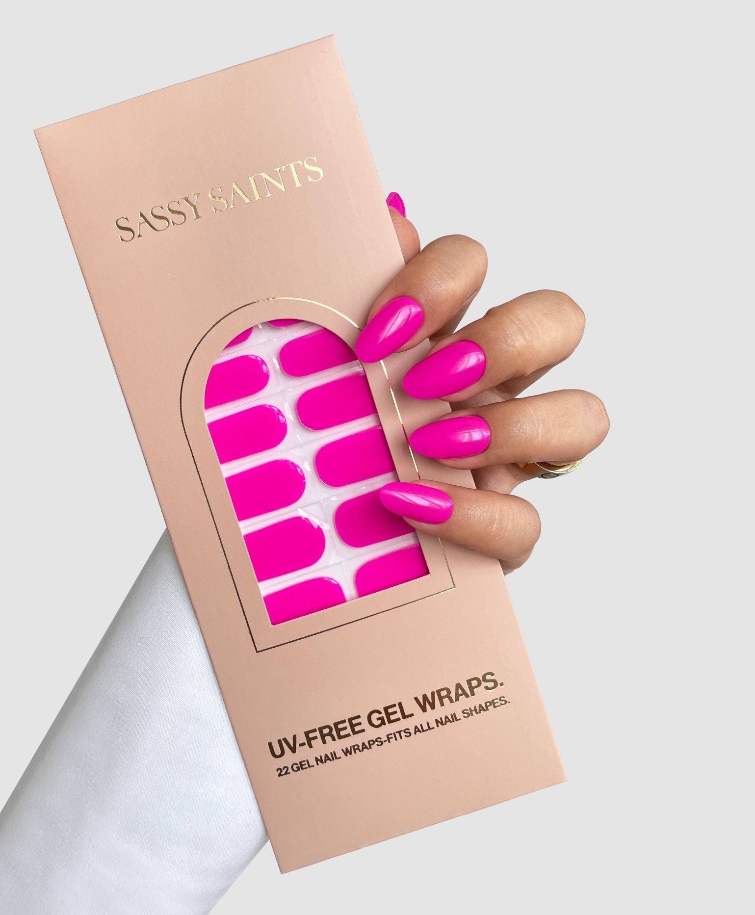 A hand with long, bright pink nails holds a peach-colored Sassy Saints - Brings The Salon Home package labeled Pink Hype Gel Wrap, containing matching pink UV-free gel nail wraps.
