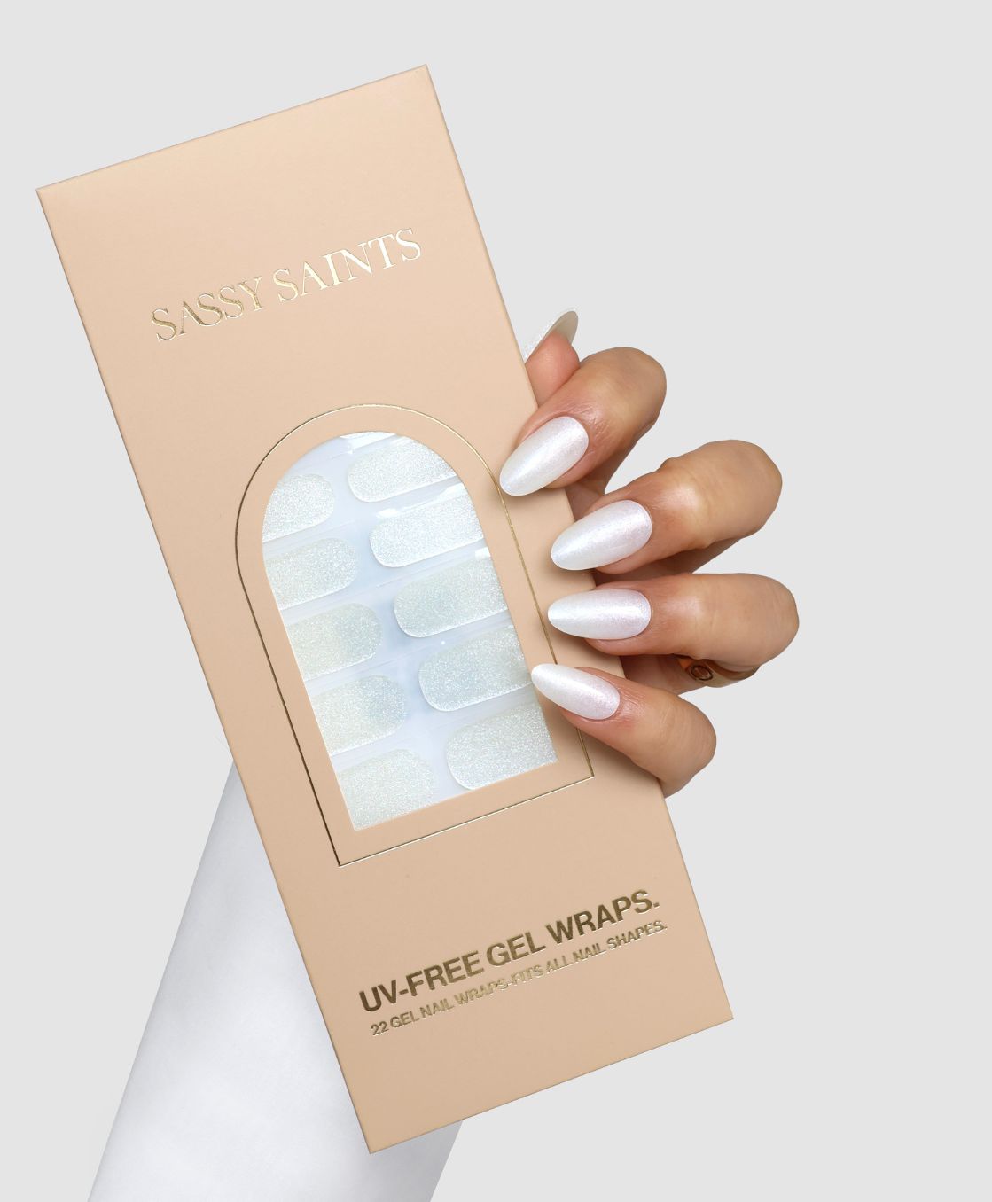 A hand with manicured, shimmery white nails holds a beige Sassy Saints – Brings The Salon Home box labeled "Icy Veil Gel Wrap," featuring UV-free gel nail wraps in shimmery white, set against a light gray background.