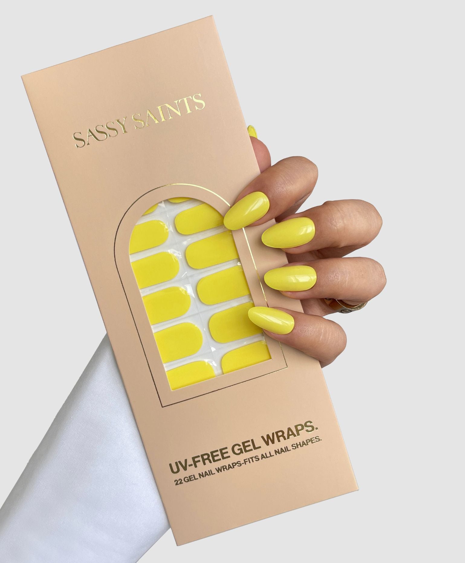 A hand with long, yellow nails holds a beige Sassy Saints - Brings The Salon Home box labeled Citrus Pop Gel Wrap, featuring 22 UV-free yellow gel nail wraps designed to fit various nail shapes.