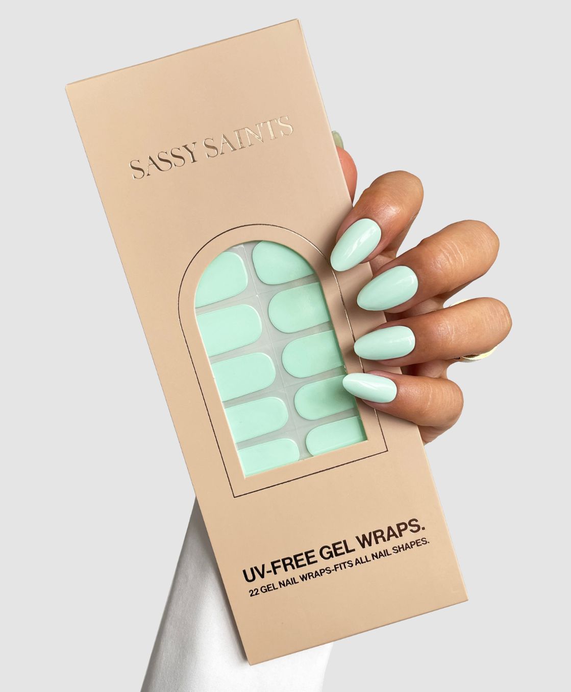 A hand with long, oval mint green nails holds a beige Sassy Saints - Brings The Salon Home package containing Mint To Be Gel Wrap UV-free nail wraps in matching mint green.
