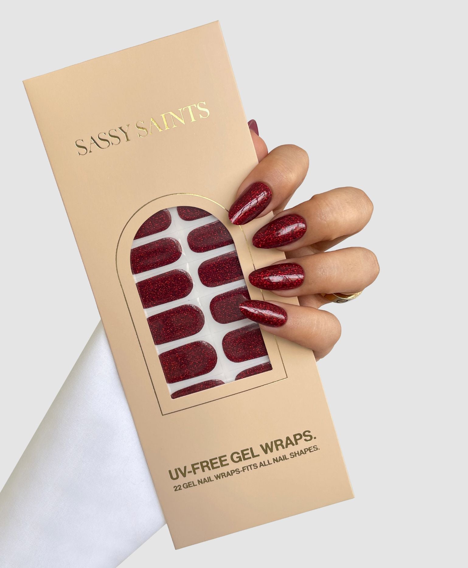 A hand with long, oval-shaped dark red glitter nails holds a beige Sassy Saints - Brings The Salon Home box containing Sparkling Ember Gel Wraps. The background is light gray.