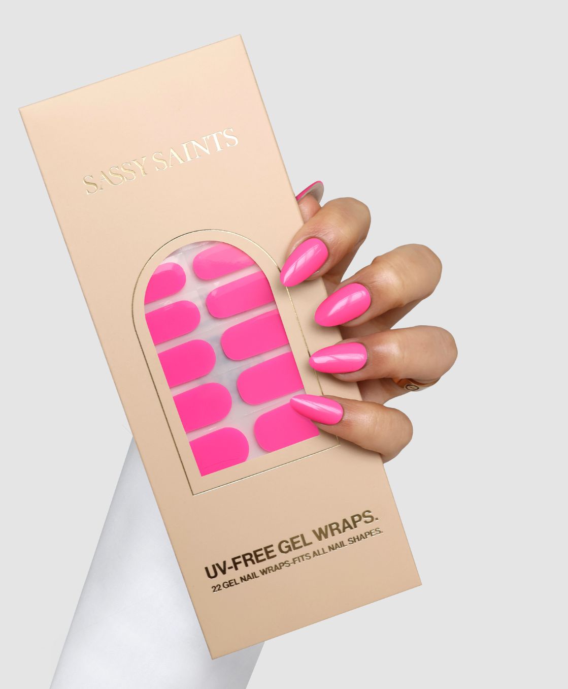 A hand with long, glossy pink nails holds a beige Sassy Saints - Brings The Salon Home box labeled Pink Problem Gel Wrap, featuring a window that displays the bright pink UV-free gel nail wraps inside.